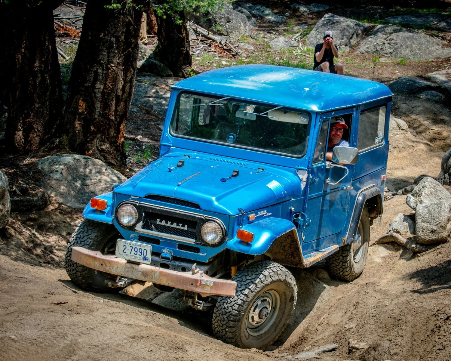 Blue hardtop Toyota FJ40 Land Cruiser climbing a sandy rock step with a spectator on the rocks above on the Ringing Rocks trail near Pipestone, Montana