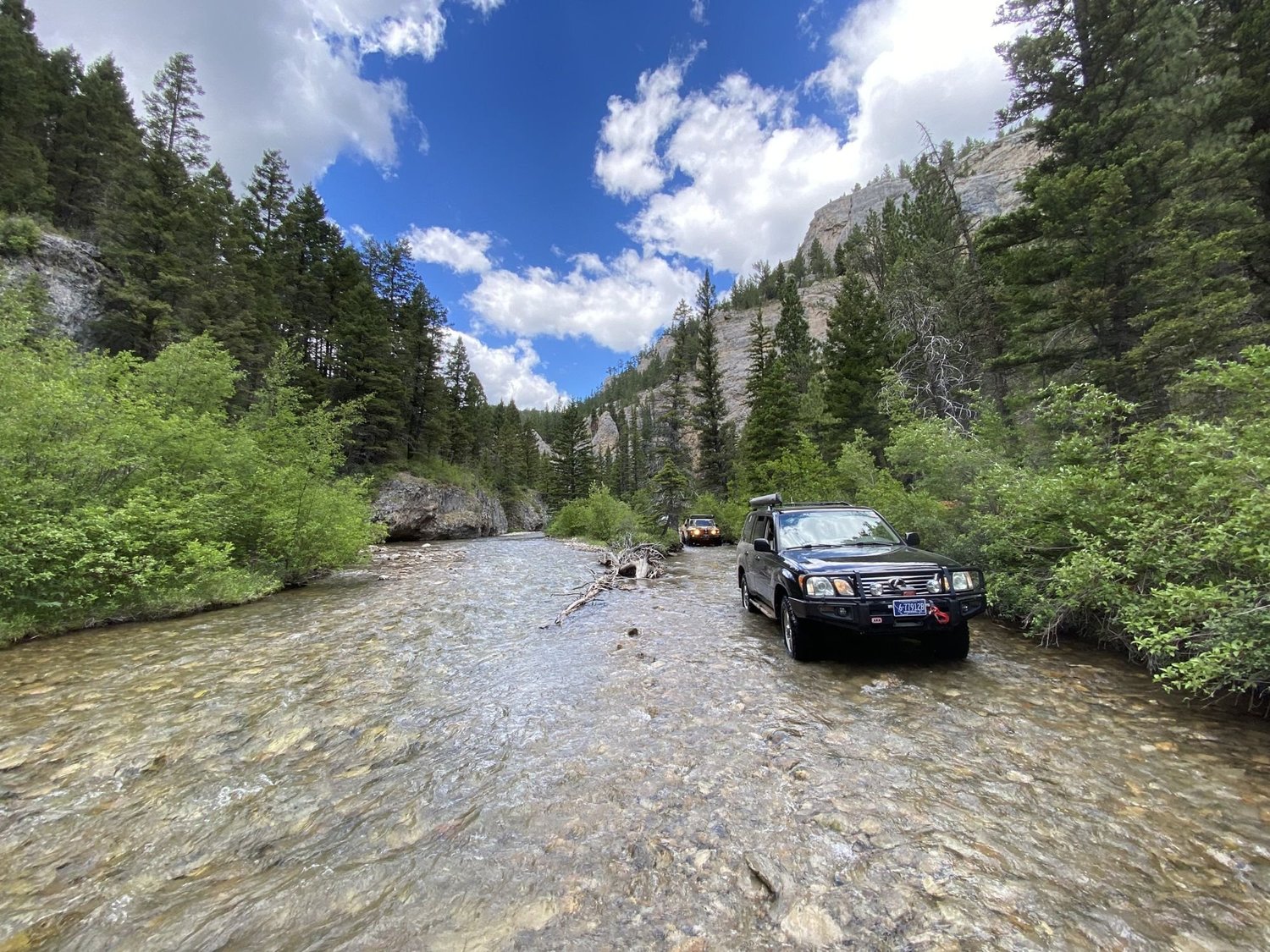 Black Lexus LX 470 crossing a wide rocky creek in a forested canyon with limestone cliffs on the Middle Fork Judith River road in the Little Belt Mountains, Montana