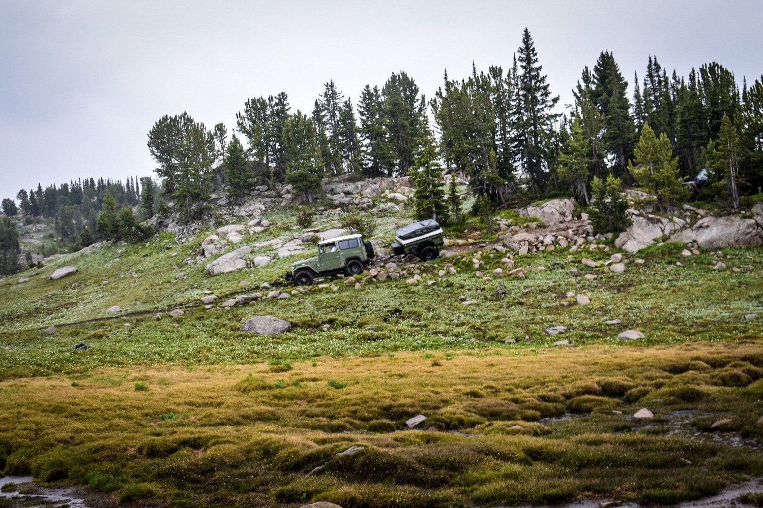 Green FJ40 and another truck crossing a boulder-strewn hillside above a golden meadow near Cooke City, Montana