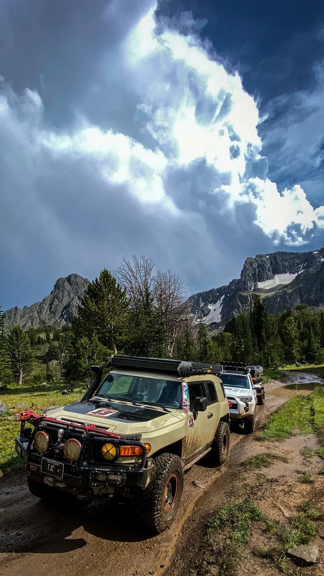 Tan Toyota FJ Cruiser leading a line of trucks through a muddy meadow under storm clouds on the Independence road above the Boulder River, Montana