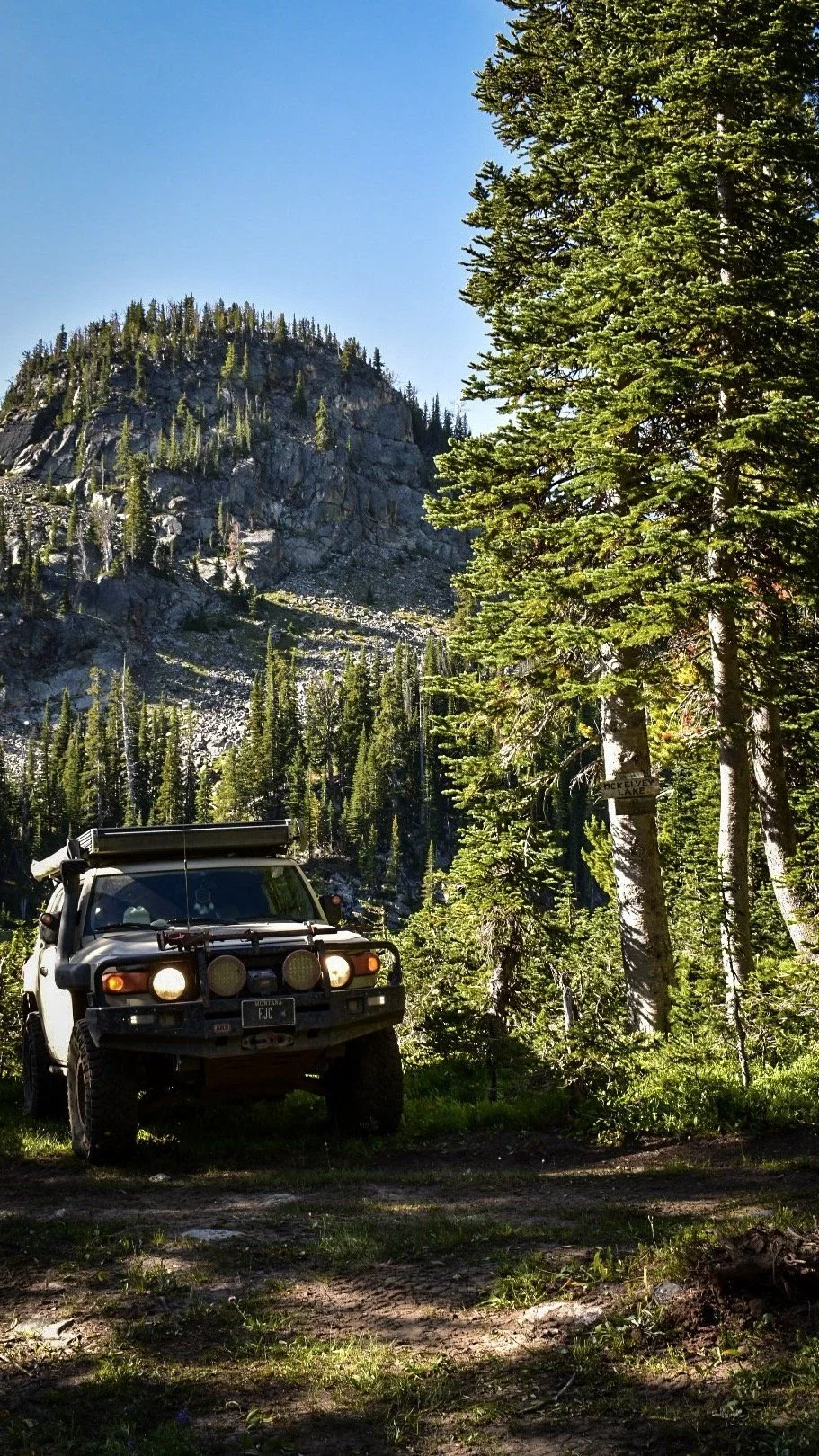Tan FJ Cruiser with lights on parked below a granite peak in the pines on the North Meadow Creek trail to McKelvey Lake in the Tobacco Root Mountains, Montana
