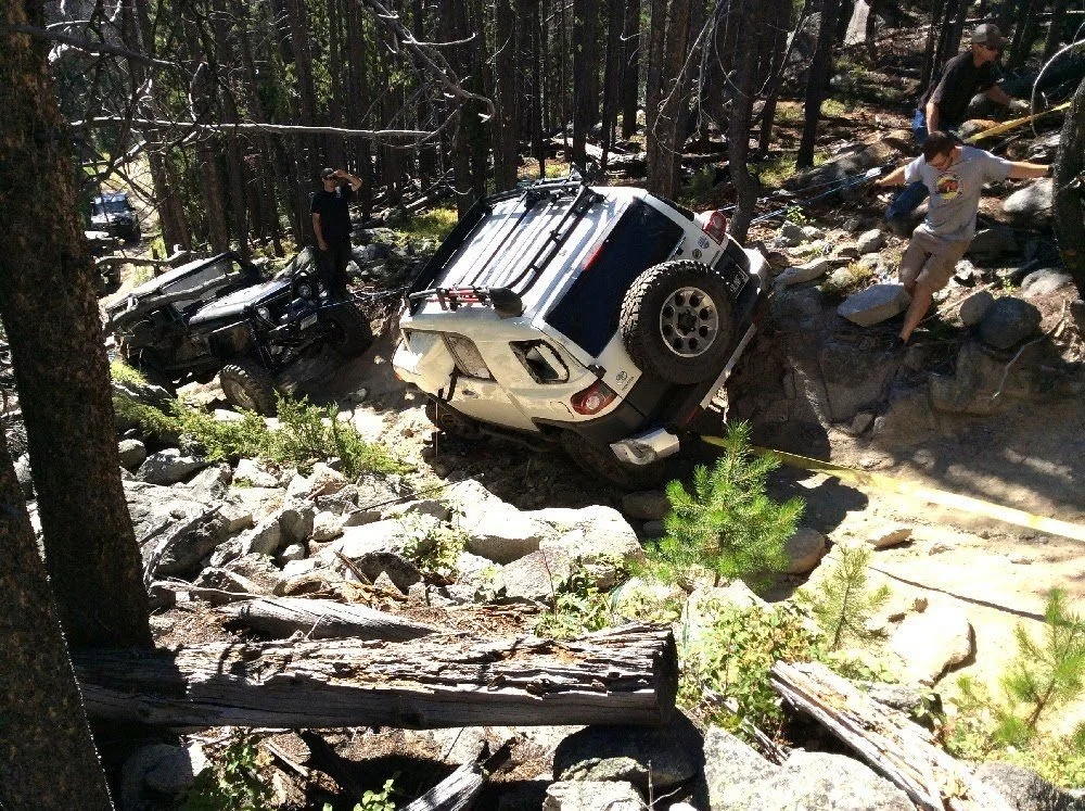 White FJ Cruiser back on its wheels in a boulder field with people standing nearby on a rocky forest trail in Montana
