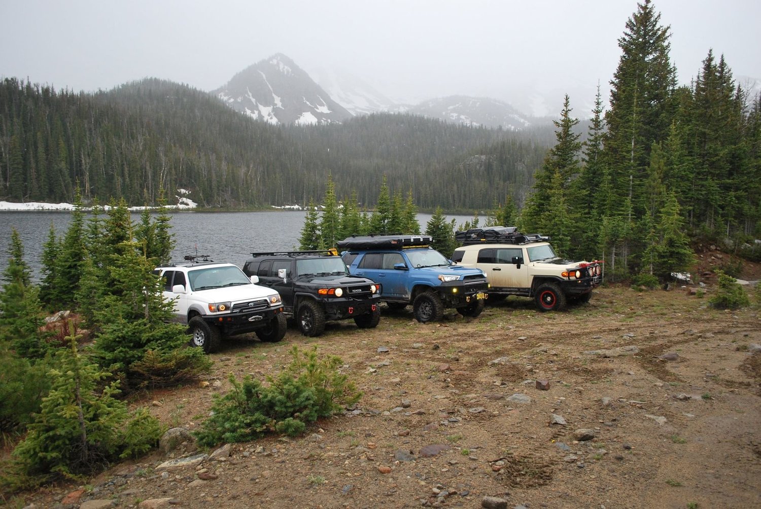 Four Toyotas lined up on the shore of a mountain lake below cloudy snow-capped peaks on the North Meadow Creek Twin Lakes trail in the Tobacco Root Mountains, Montana