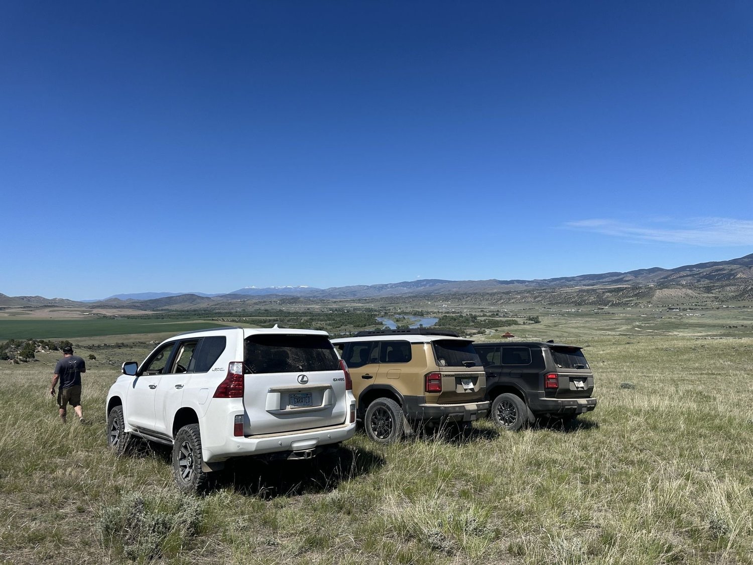 White Lexus GX, tan Land Cruiser 250 and a dark truck parked on a grassy hill above a valley at XOverland Ranch near Bozeman, Montana