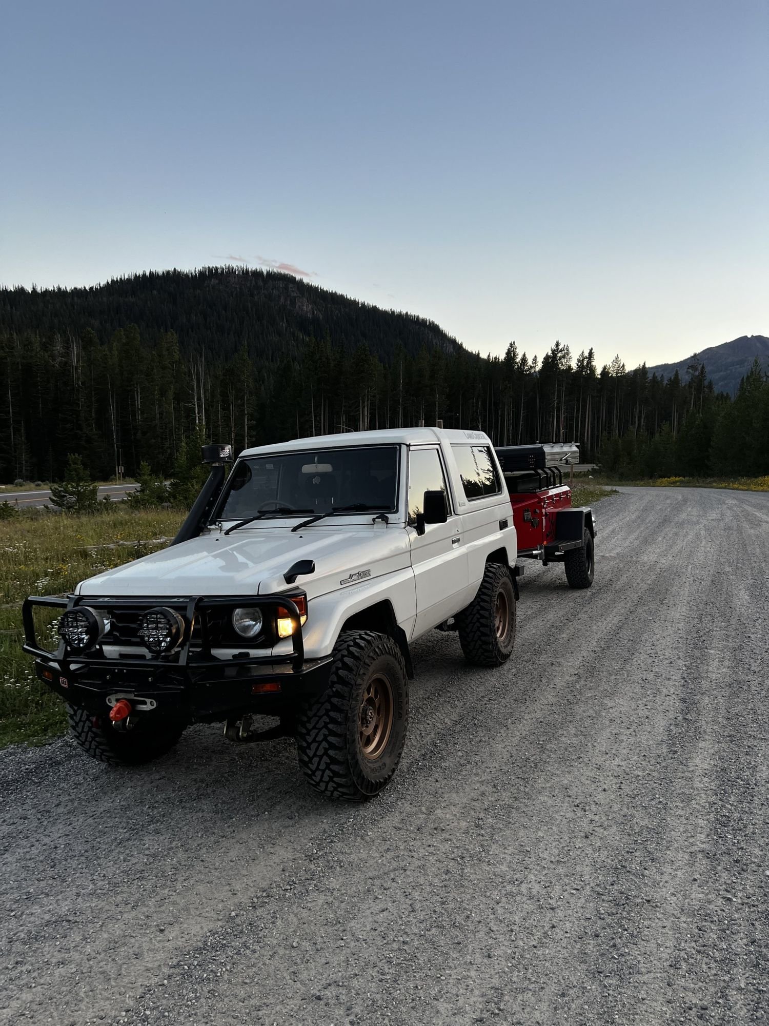 White short-wheelbase Toyota Land Cruiser 70 Series towing a small red trailer on a gravel road near Cooke City, Montana