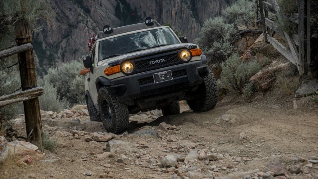 Tan FJ Cruiser with amber lights crawling over rocks past a wooden fence in the timber on the Morrison Jeep Trail in Clarks Fork Canyon near Cody, Wyoming
