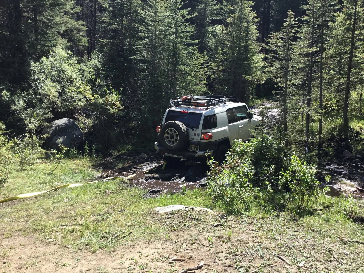 White FJ Cruiser stuck in a mud bog in a clearing with a recovery strap stretched out on a rocky forest trail in Montana