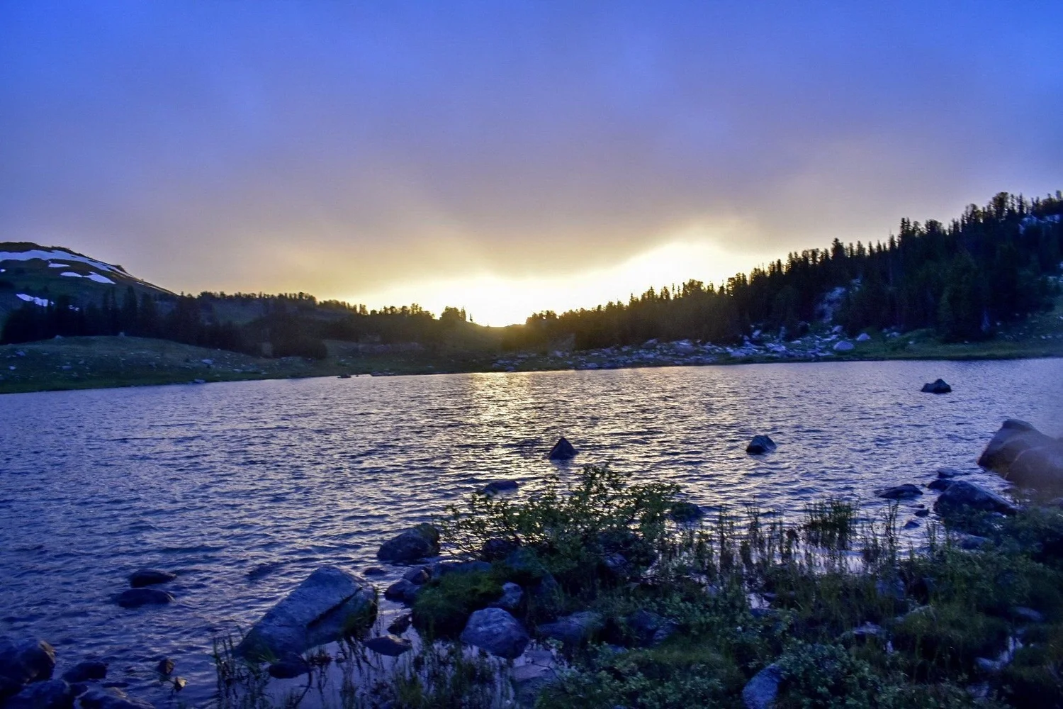 Orange sunset glowing behind dark timber over a rippling alpine lake near Cooke City, Montana