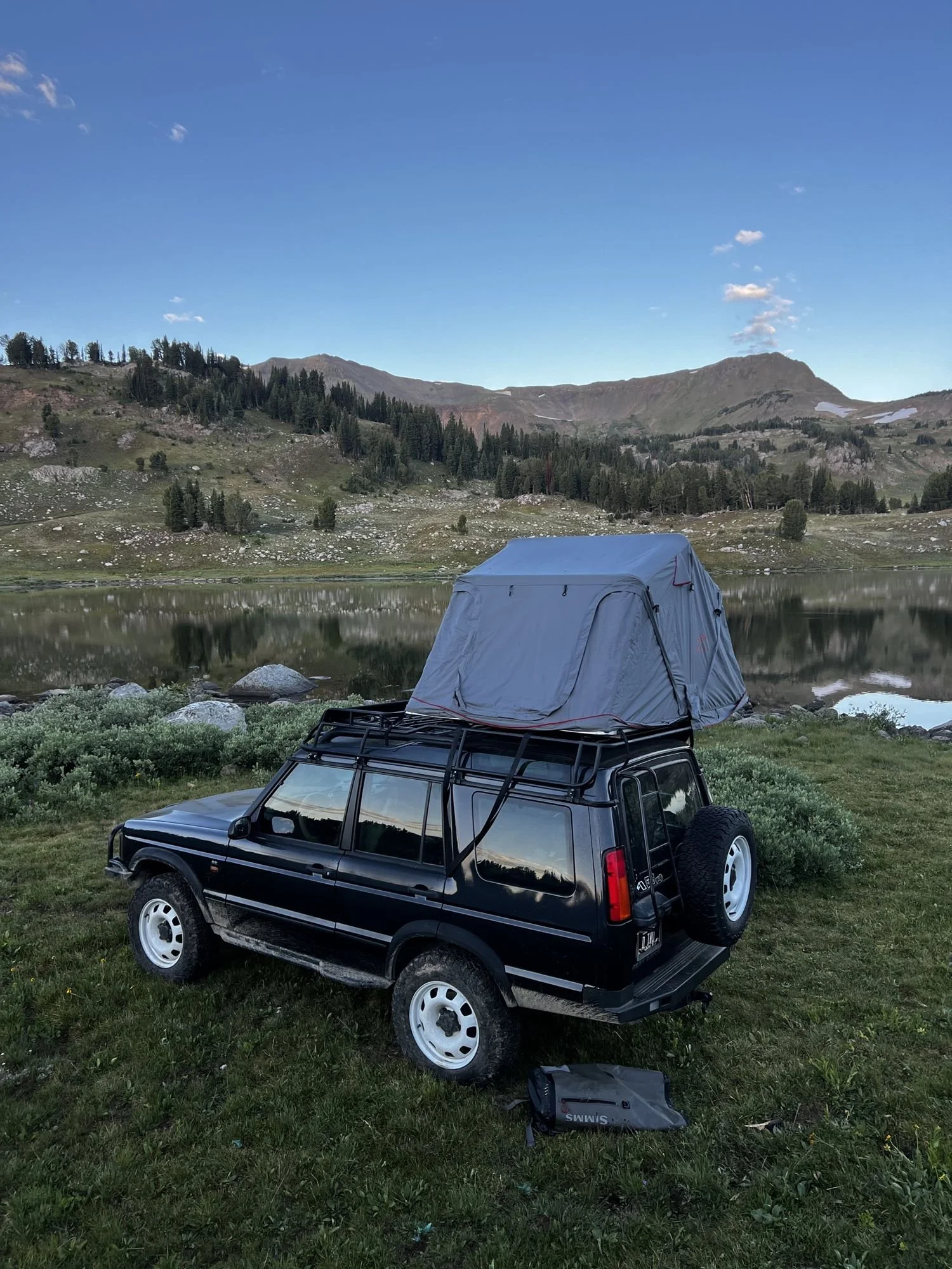 Black Land Rover Discovery with a gray rooftop tent parked beside a high lake near Cooke City, Montana