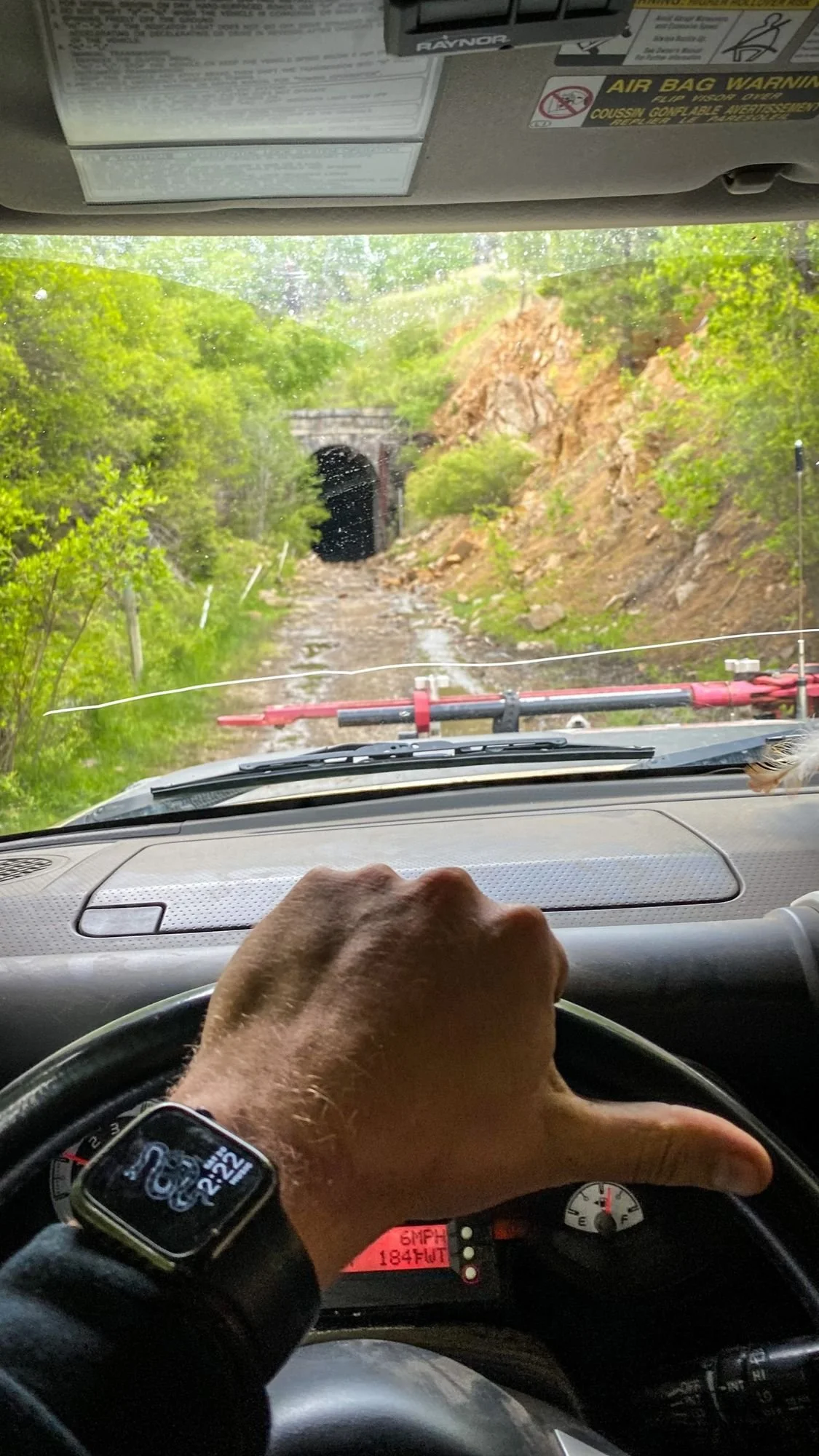 View through a truck windshield up a green, brushy grade toward the stone portal of the Wickes tunnel at the Wickes railroad tunnel near Jefferson City, Montana