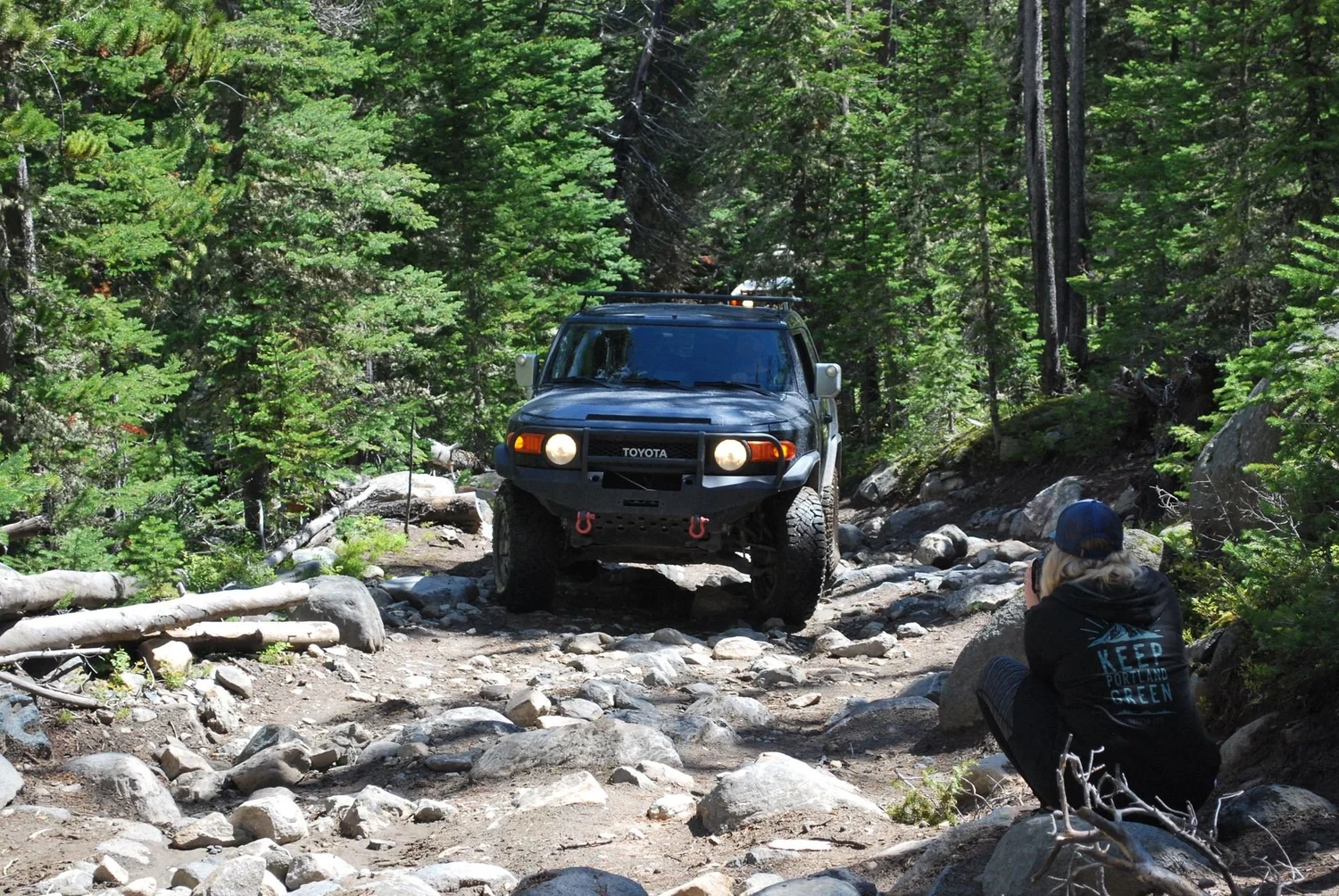 Dark Toyota coming down a rocky section toward a spotter crouched on the rocks on the North Meadow Creek Twin Lakes trail in the Tobacco Root Mountains, Montana
