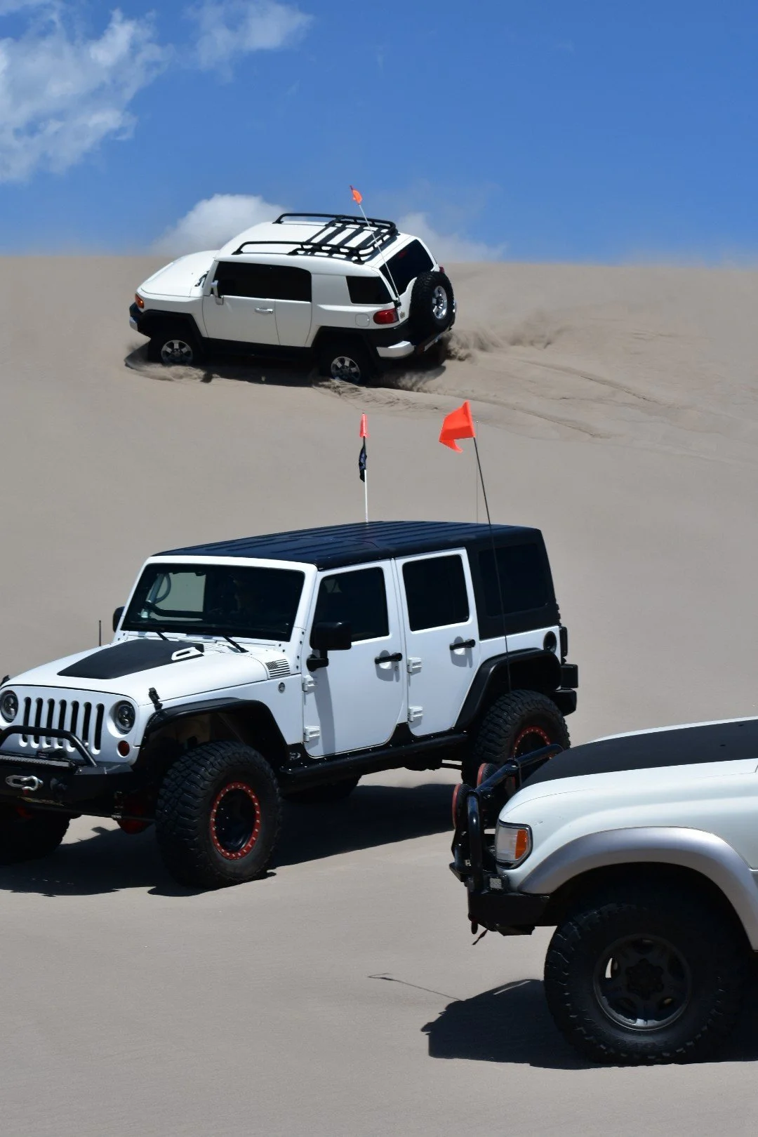 White Toyota FJ Cruiser cresting a dune lip with a flagged white Jeep Wrangler in the foreground at the St. Anthony Sand Dunes in Idaho