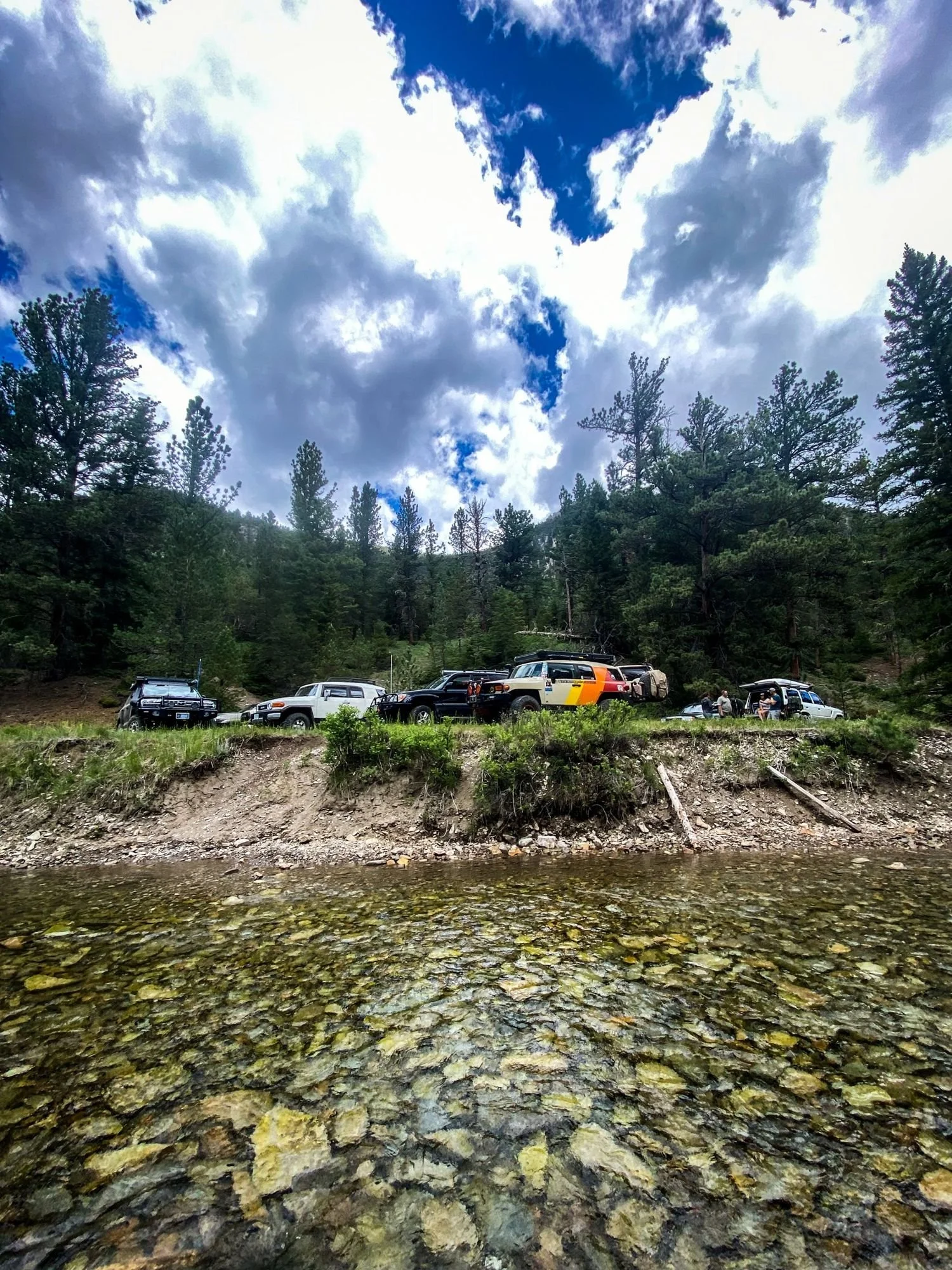 Line of trucks parked across a clear river with smooth stones in the foreground on the Middle Fork Judith River road in the Little Belt Mountains, Montana