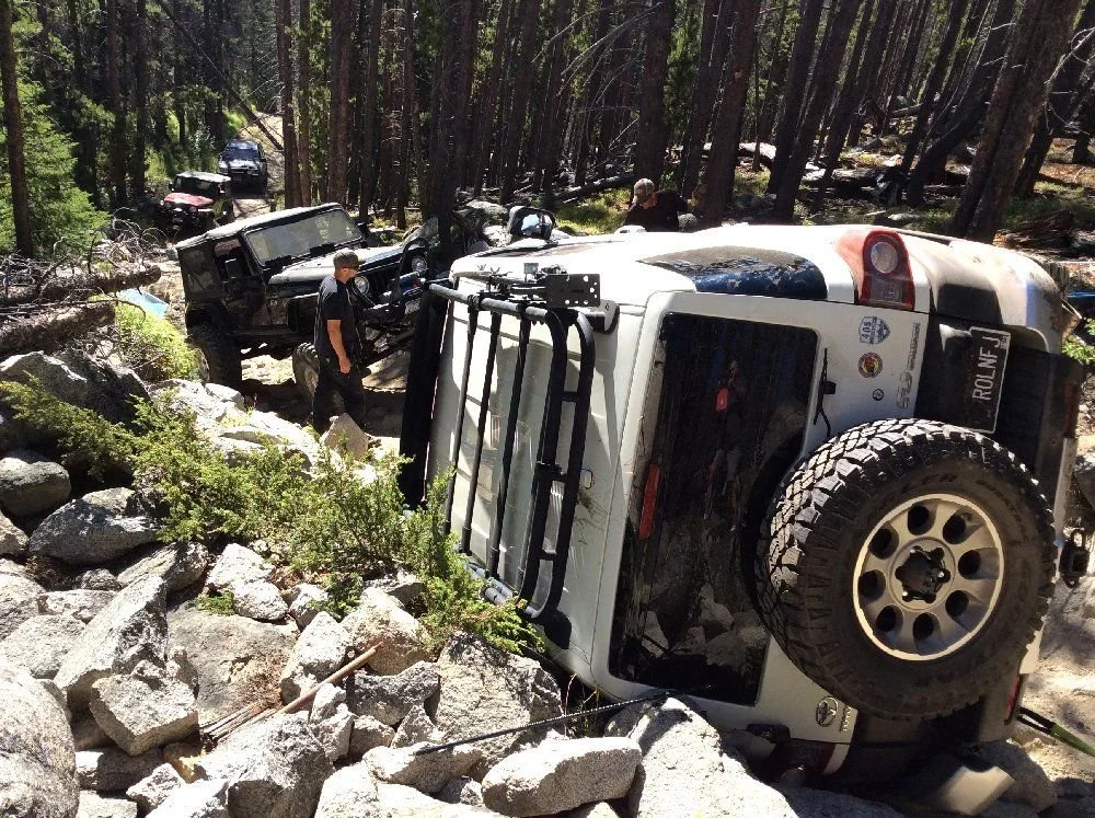 White FJ Cruiser on its side with straps attached and people working uphill on a rocky forest trail in Montana