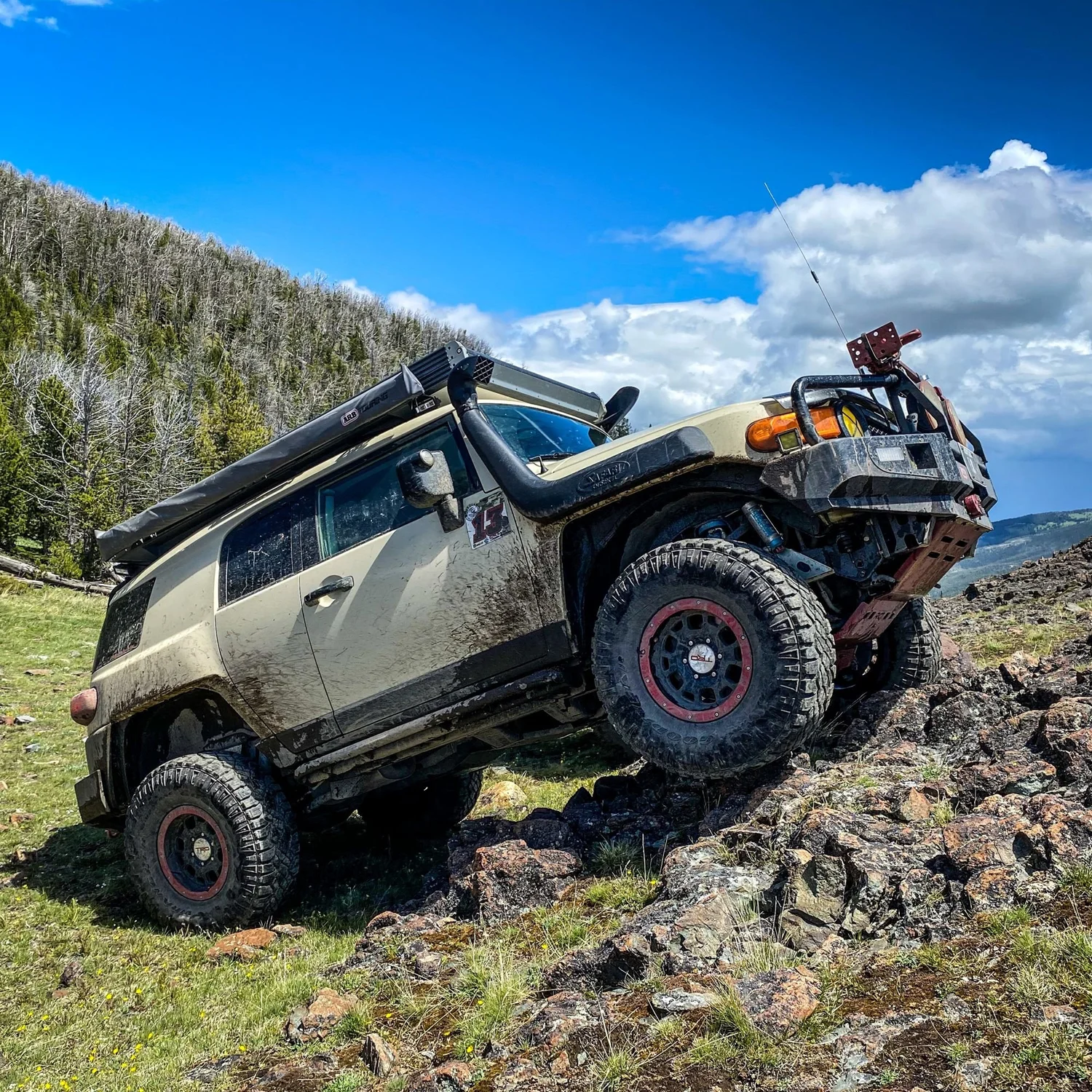 Tan Toyota FJ Cruiser with red wheels and a snorkel climbing a rock outcrop on a ridge under a blue sky in the Elkhorn Mountains near Boulder, Montana