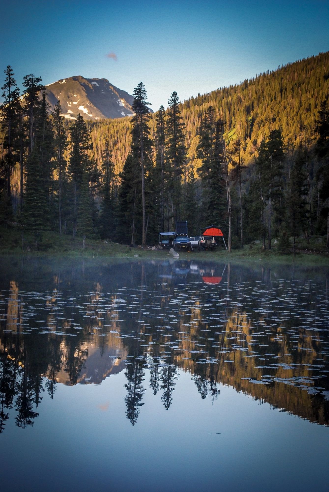Peak lit orange at sunrise above a dark forest and a calm lake with a camp on the far shore on the North Meadow Creek Twin Lakes trail in the Tobacco Root Mountains, Montana