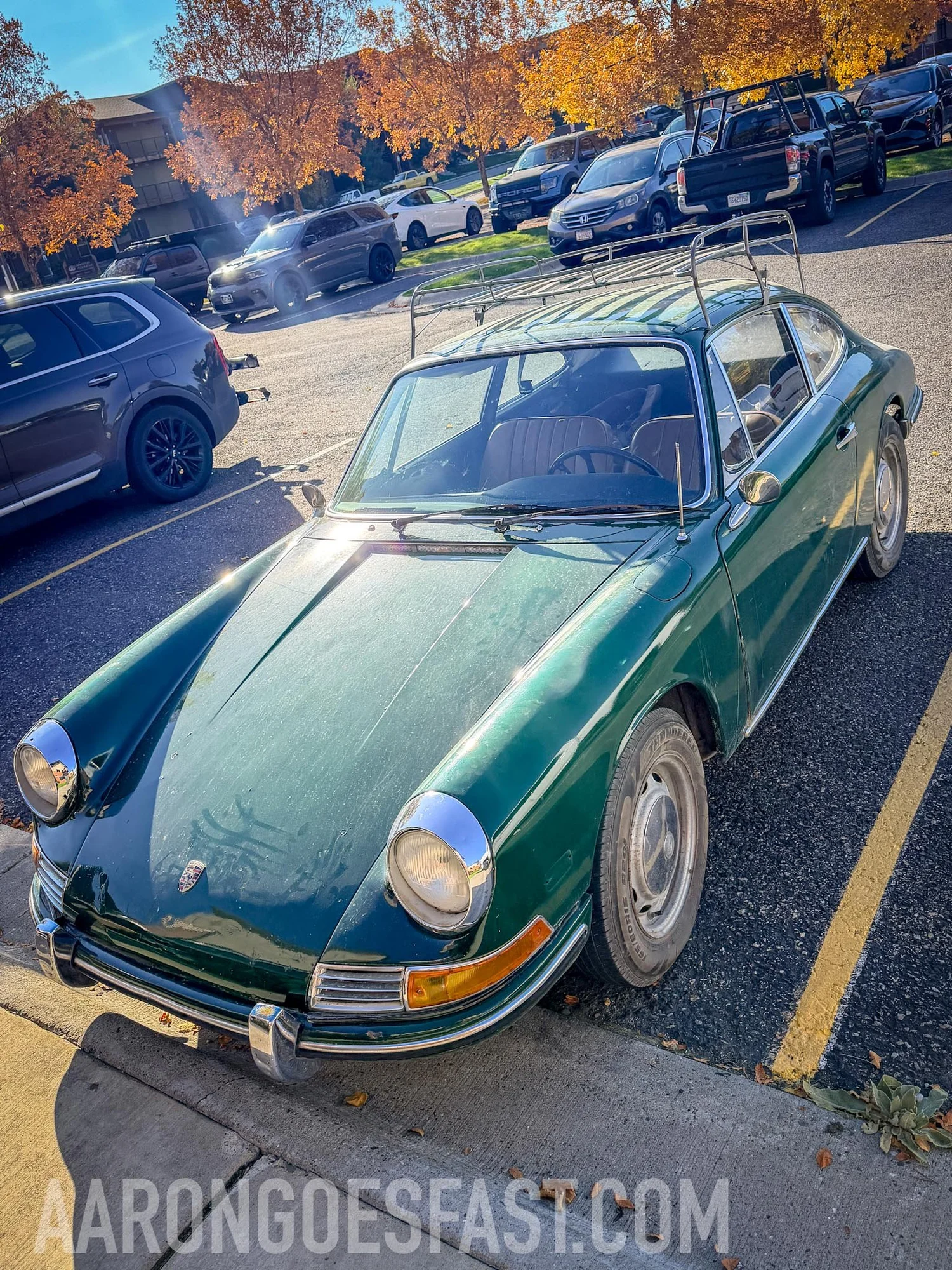 Front three-quarter view of a green early Porsche 912 with round headlights, amber turn signals and a roof rack, fall trees behind