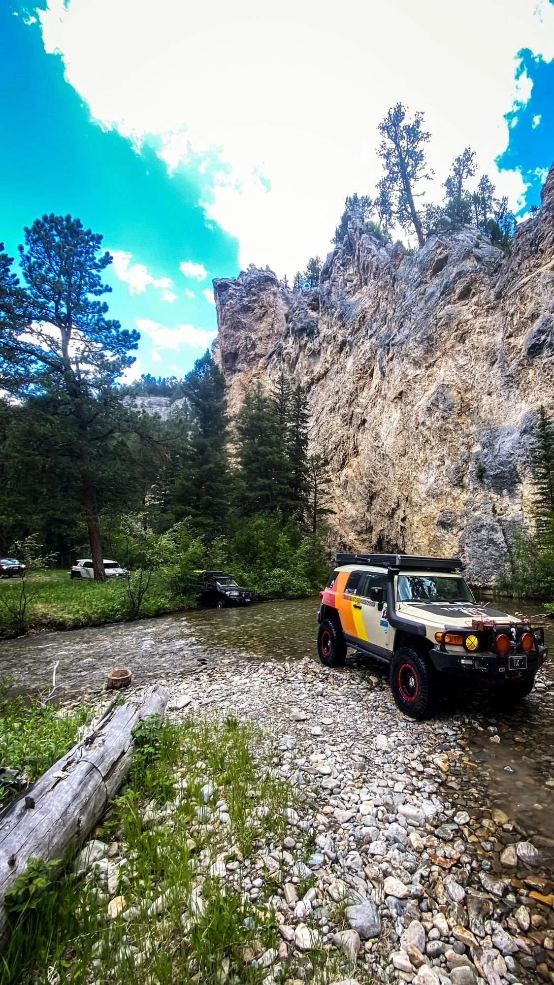 FJ Cruiser with orange stripes crossing a shallow rocky creek below a tall limestone cliff on the Middle Fork Judith River road in the Little Belt Mountains, Montana
