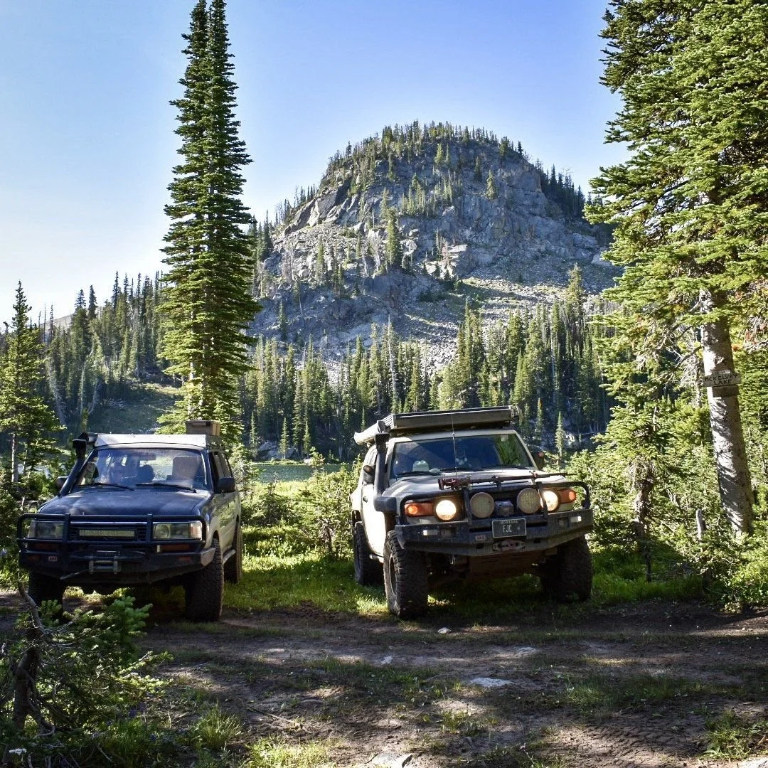 Johnny’s blue Land Cruiser 80 Series and a tan FJ Cruiser with lights on parked in the timber below a granite peak on the North Meadow Creek trail to McKelvey Lake in the Tobacco Root Mountains, Montana