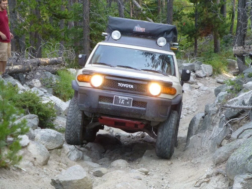 Tan FJ Cruiser with lights on crawling down a boulder trail in the pines on a rocky forest trail in Montana