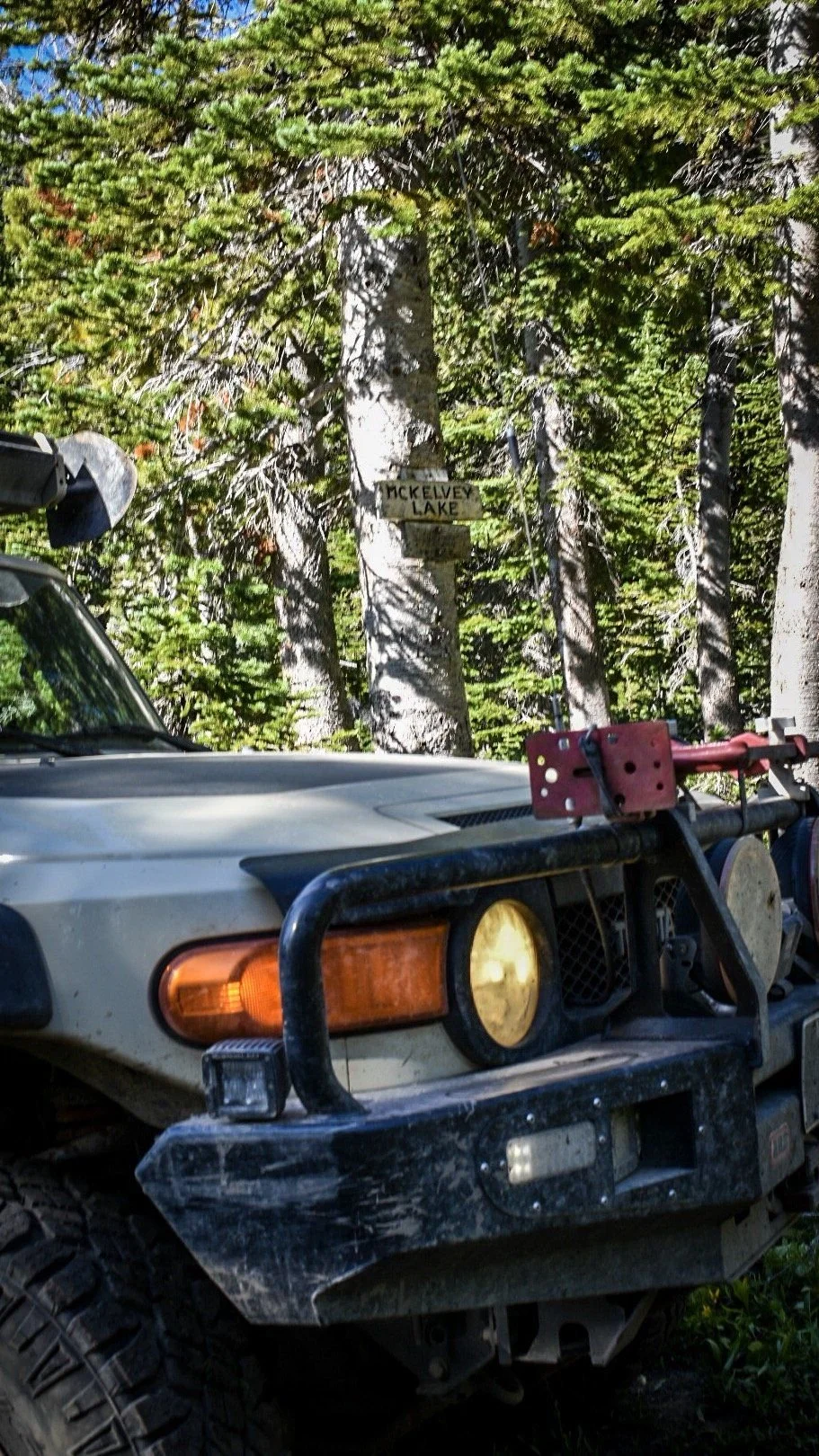 Front corner of a tan FJ Cruiser beside a hand-carved McKelvey Lake sign on a tree on the North Meadow Creek trail to McKelvey Lake in the Tobacco Root Mountains, Montana