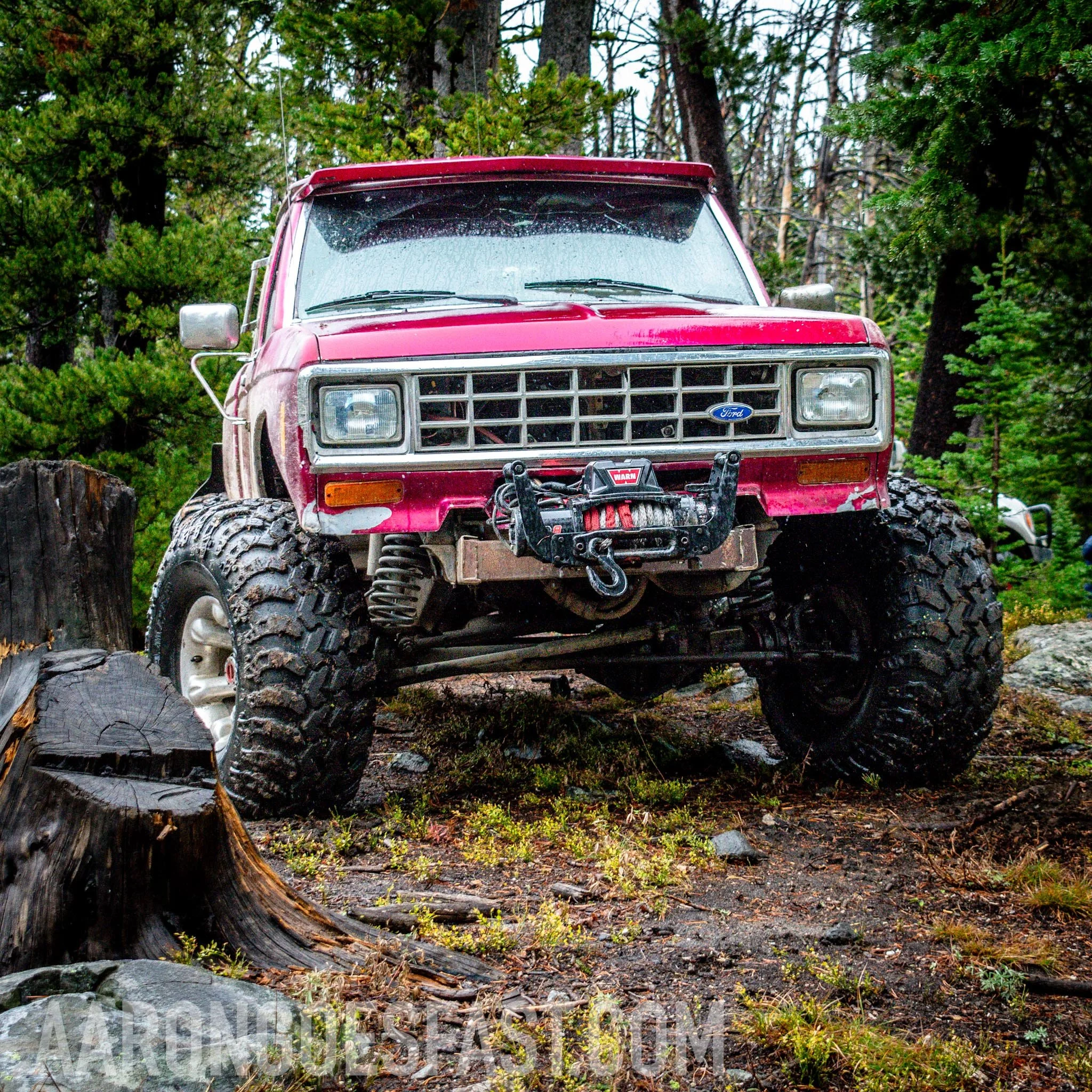 Red vintage Ford Bronco climbing over a stump, front axle fully articulated