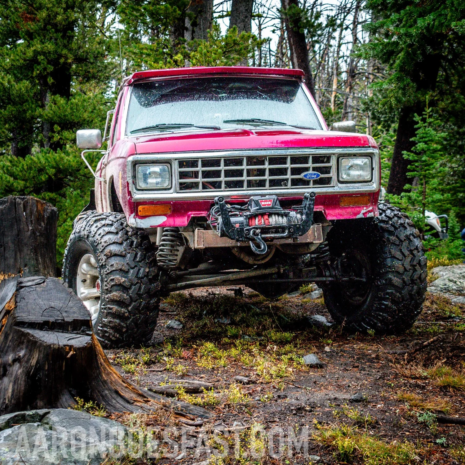 Red vintage Ford Bronco climbing over a stump, front axle fully articulated
