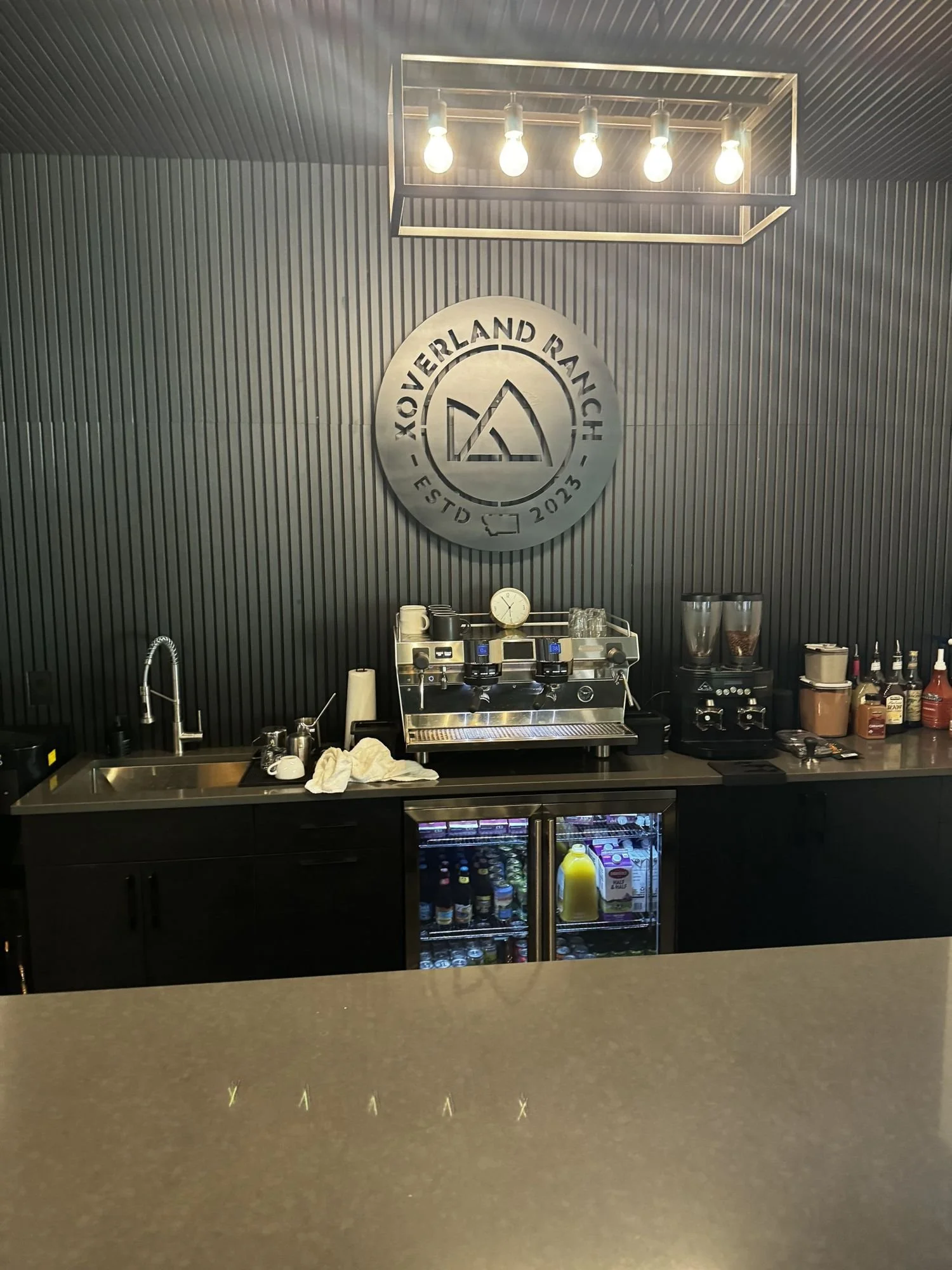 Coffee bar with an espresso machine below a round XOverland Ranch logo on a dark wall