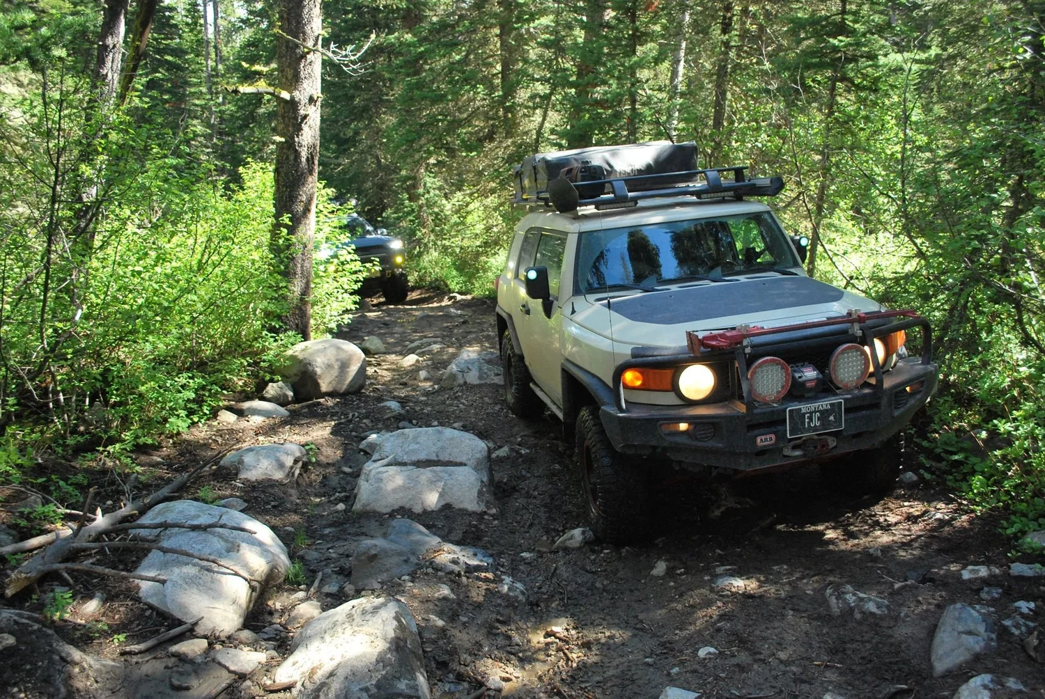 Tan FJ Cruiser with lights on picking through boulders on a rocky trail in the timber on the North Meadow Creek Twin Lakes trail in the Tobacco Root Mountains, Montana