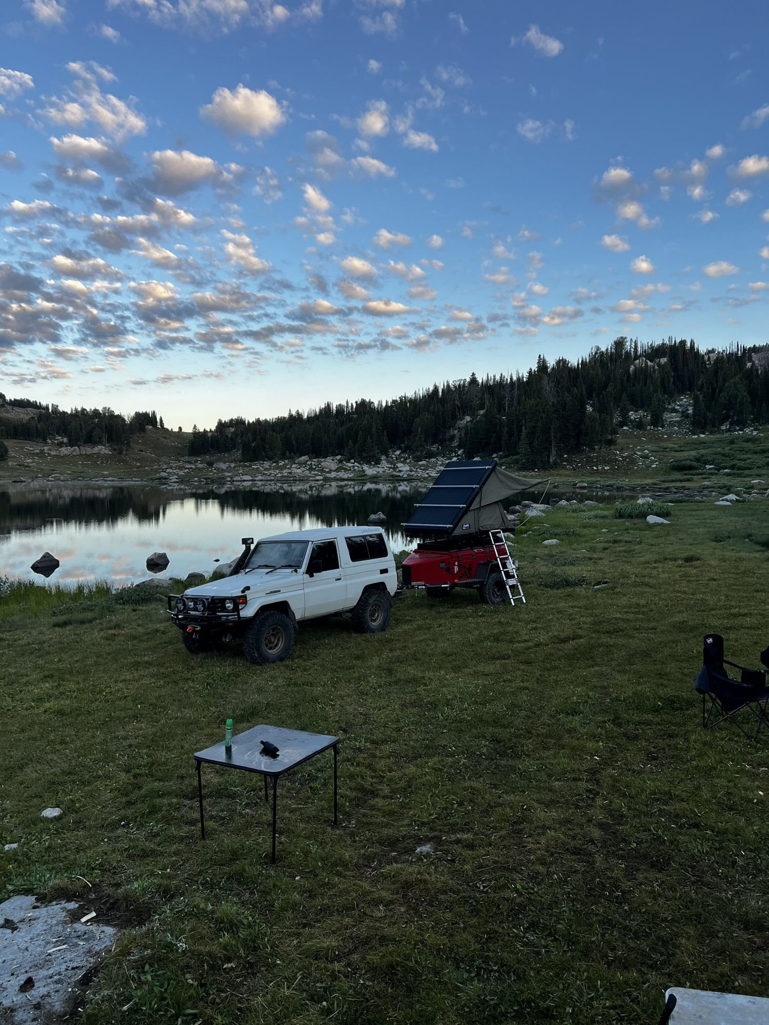 Camp in a meadow beside a lake with a white truck, a rooftop tent and a camp table under a cloudy sky near Cooke City, Montana