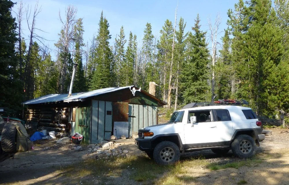 White FJ Cruiser parked by an old cabin in the pines with a tan FJ Cruiser behind on a rocky forest trail in Montana