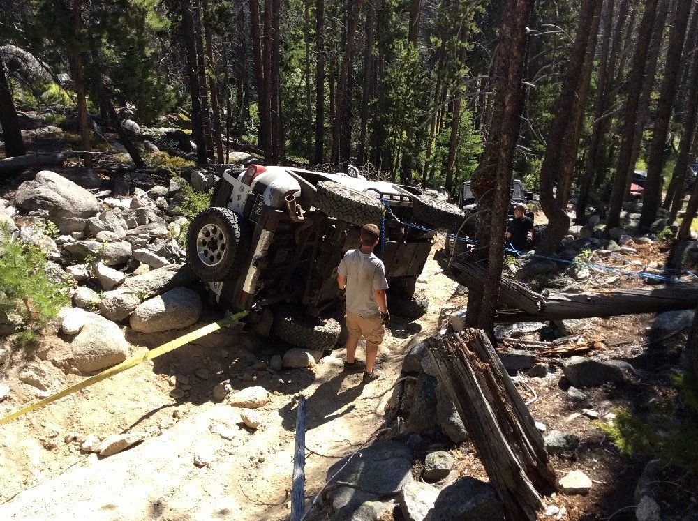 White FJ Cruiser on its side among boulders on a sandy forest trail with a person standing nearby on a rocky forest trail in Montana