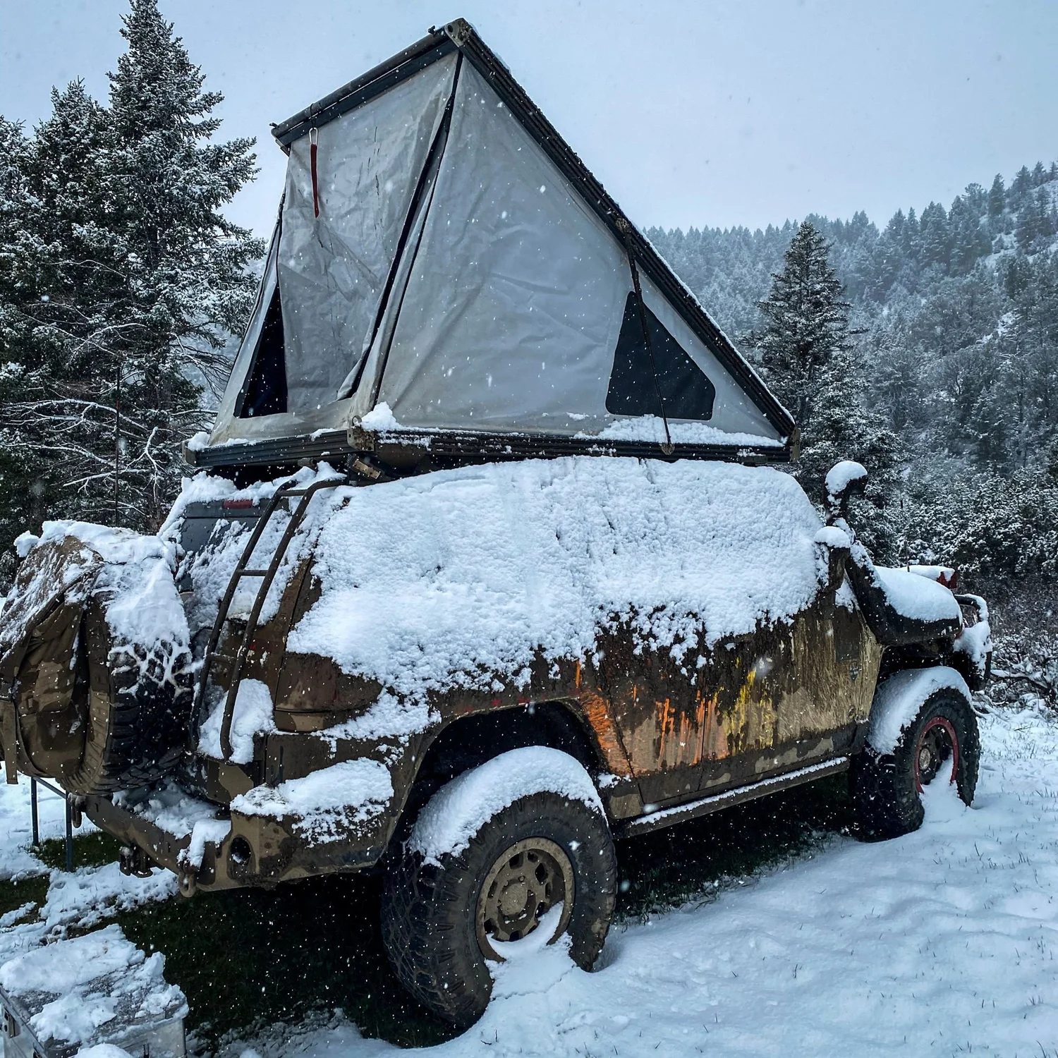 Mud-caked truck with a hard-shell rooftop tent covered in fresh snow among pine trees on a 406 Cruisers run in the upper Ruby River country, Montana