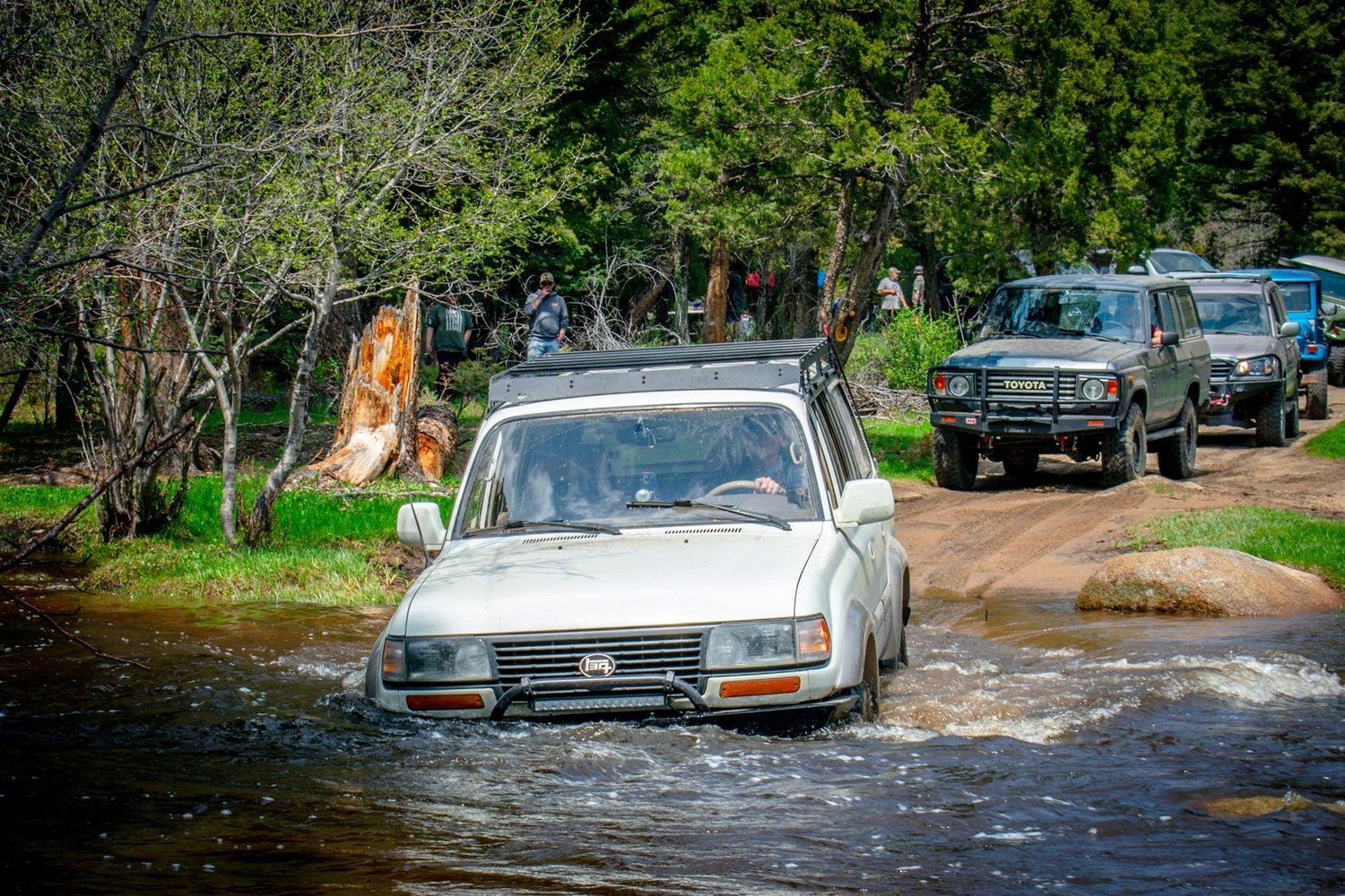 White Toyota Land Cruiser with a roof rack pushing through a creek, a green Land Cruiser waiting behind on the Ringing Rocks trail near Pipestone, Montana