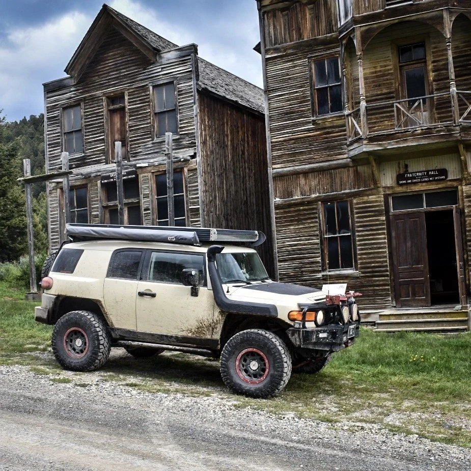 Tan Toyota FJ Cruiser with red wheels parked in front of two weathered wooden halls in the Elkhorn ghost town near Boulder, Montana