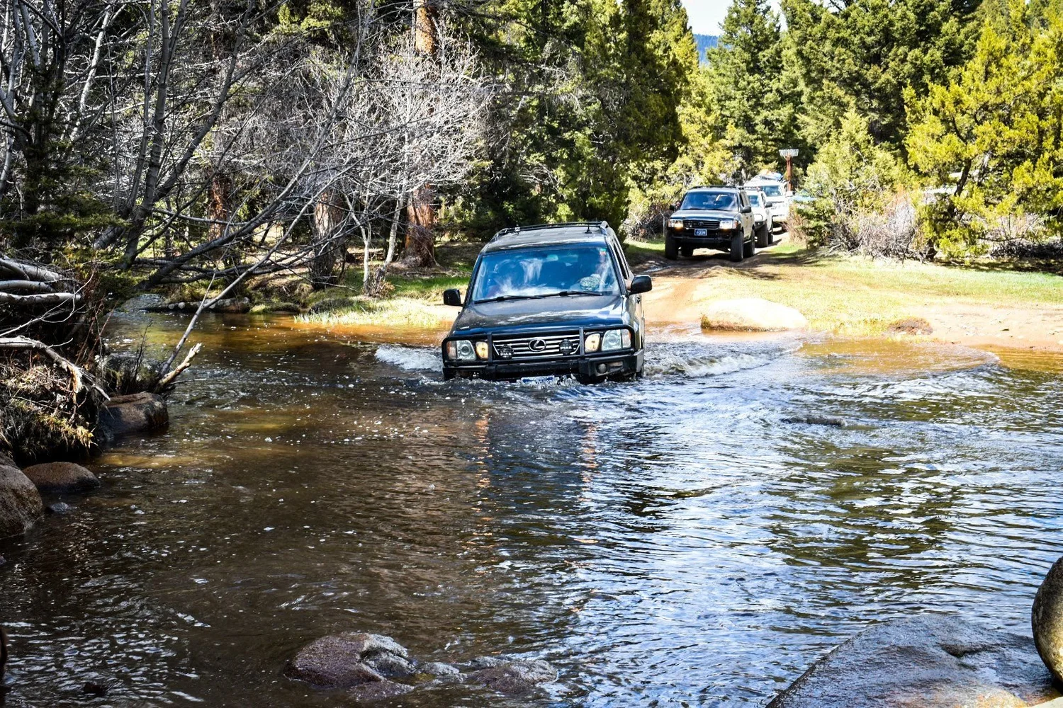 Dark Toyota SUV fording a wide clear creek with more rigs waiting on the far bank on the Ringing Rocks trail near Pipestone, Montana