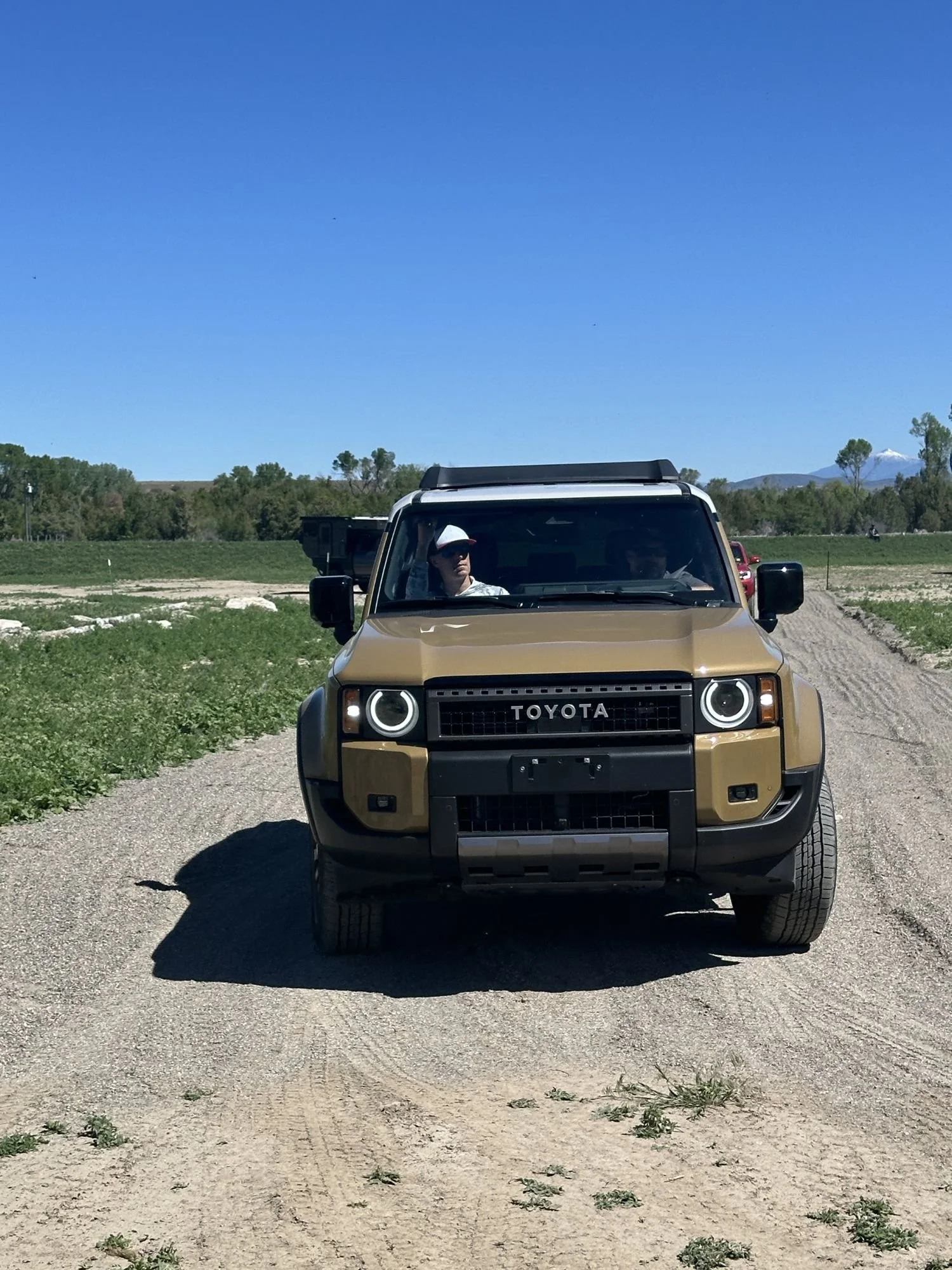 Tan 2024 Toyota Land Cruiser 250 with round headlights and a white roof driving toward the camera on a dirt course at XOverland Ranch near Bozeman, Montana