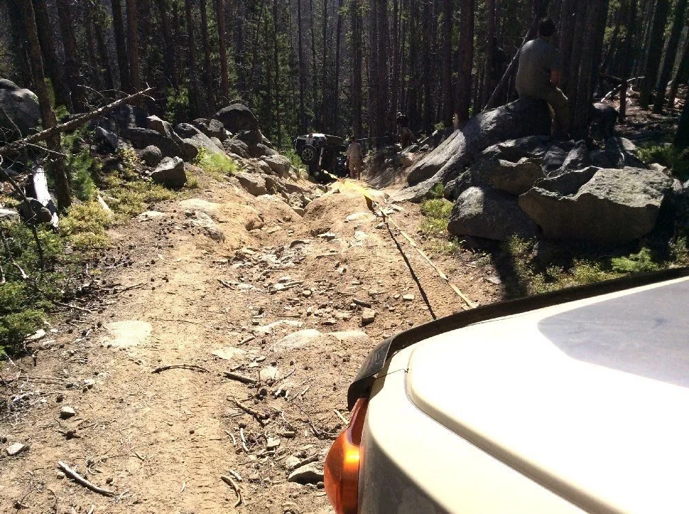 View over the hood of a tan FJ Cruiser of a recovery strap running down a sandy trail toward the rollover on a rocky forest trail in Montana