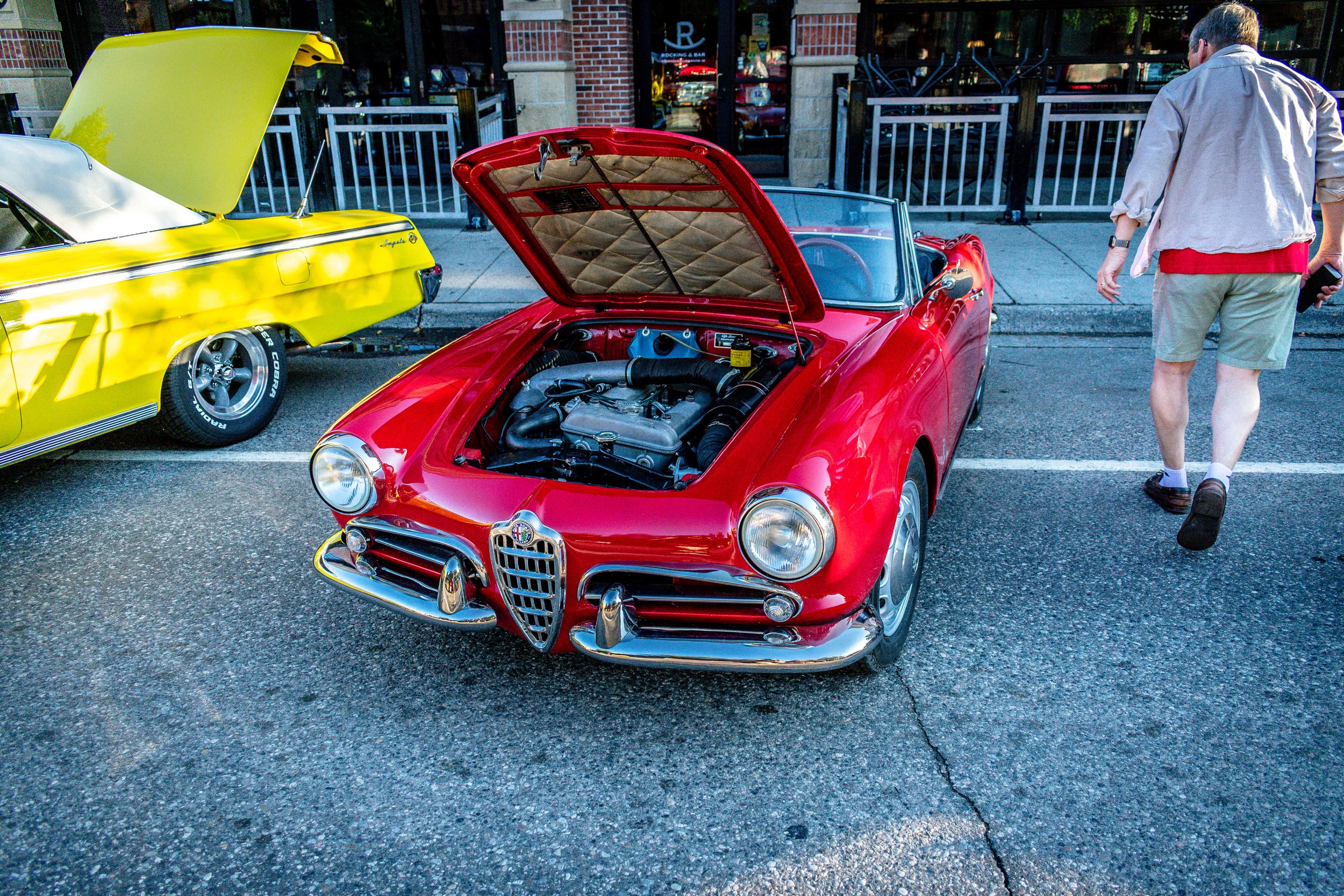 Front three-quarter view of the red Alfa Romeo Spider with the hood open, a man walking past on the sidewalk