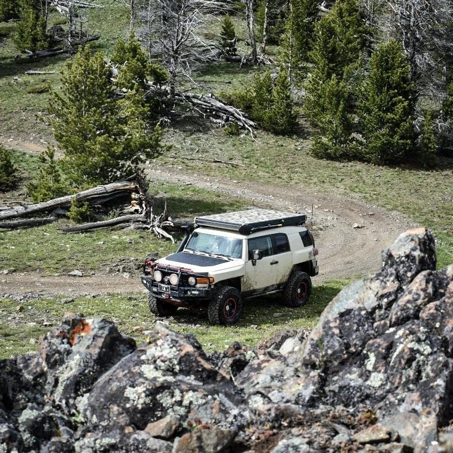Tan FJ Cruiser on a dirt trail seen past lichen-covered rocks in the Elkhorn Mountains near Boulder, Montana