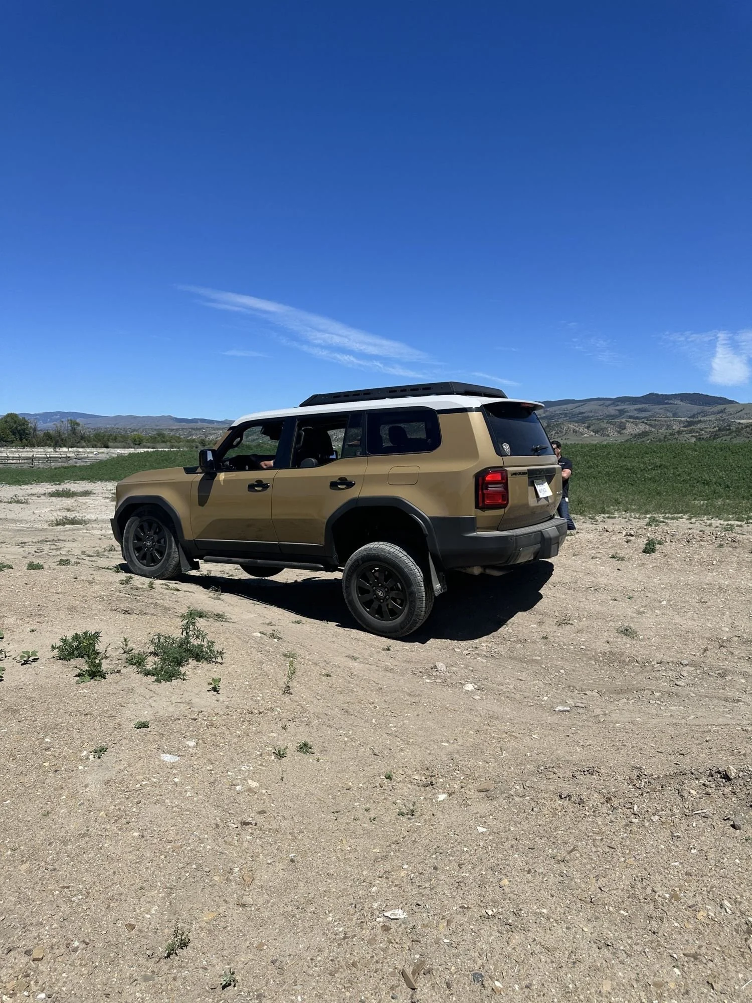 Rear three-quarter view of a tan Land Cruiser 250 with a white roof on a dirt slope at XOverland Ranch near Bozeman, Montana