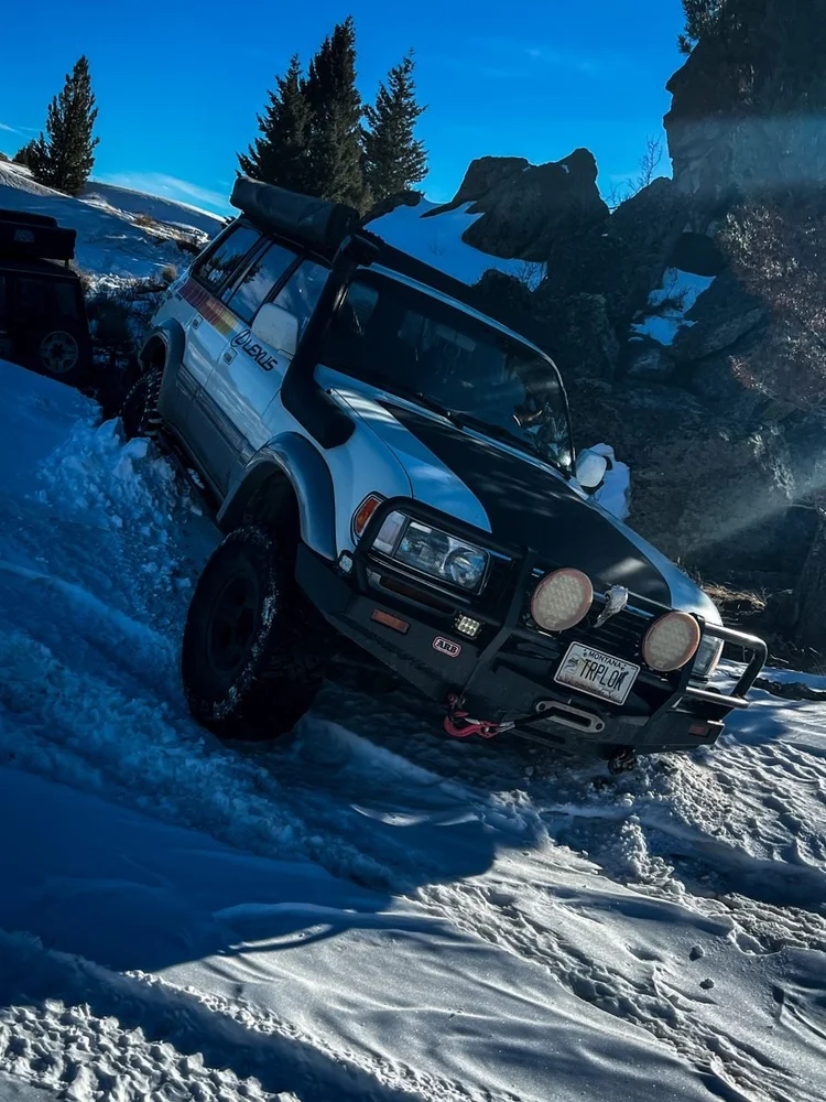 A silver Lexus LX450 with TRPLOK plates nosed into deep snow on a tilted mountain track, rocks and pines behind
