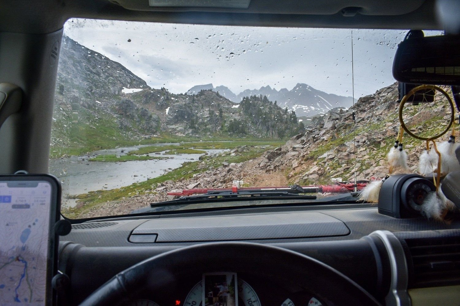 View through a windshield of a creek and lake below rocky peaks near Cooke City, Montana
