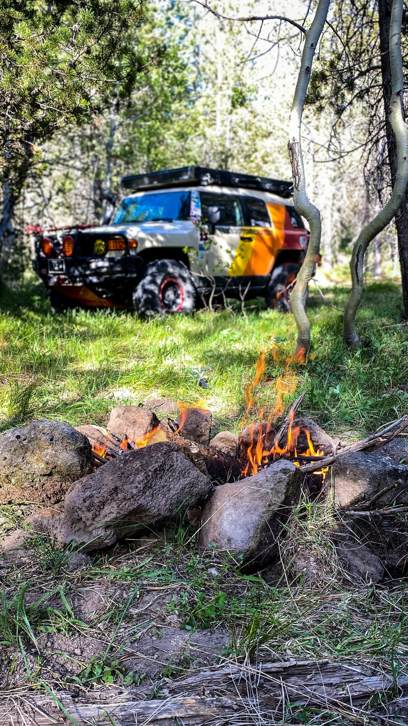 Tan Toyota FJ Cruiser with retro orange and yellow stripes and a rooftop tent parked in aspens behind a campfire