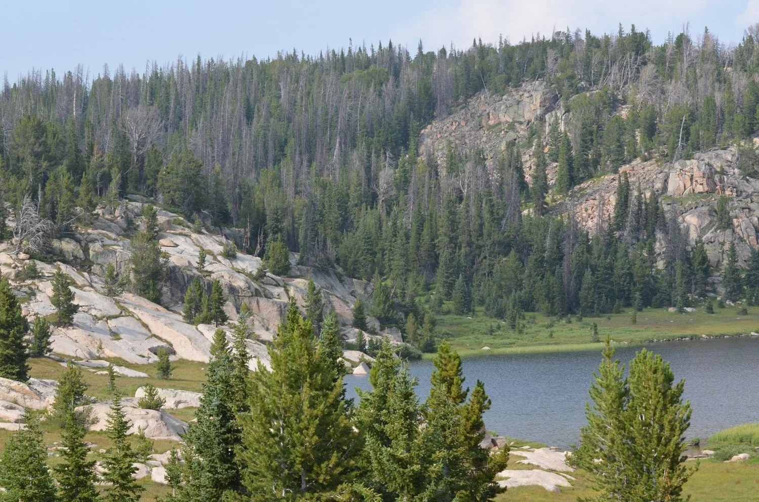 Alpine lake ringed by granite slabs and pine forest under a hazy sky on the Morrison Jeep Trail in Clarks Fork Canyon near Cody, Wyoming