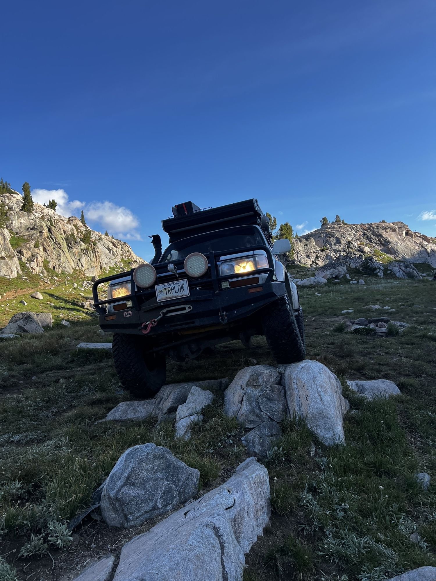 Dark Land Cruiser with lights perched on granite boulders with rocky peaks behind near Cooke City, Montana