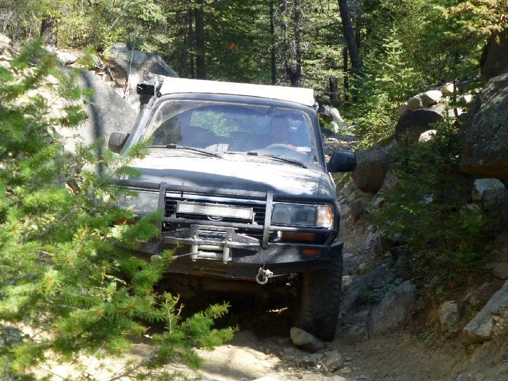 Dark gray Land Cruiser 80 Series with a winch bumper squeezing between boulders on a rocky forest trail in Montana