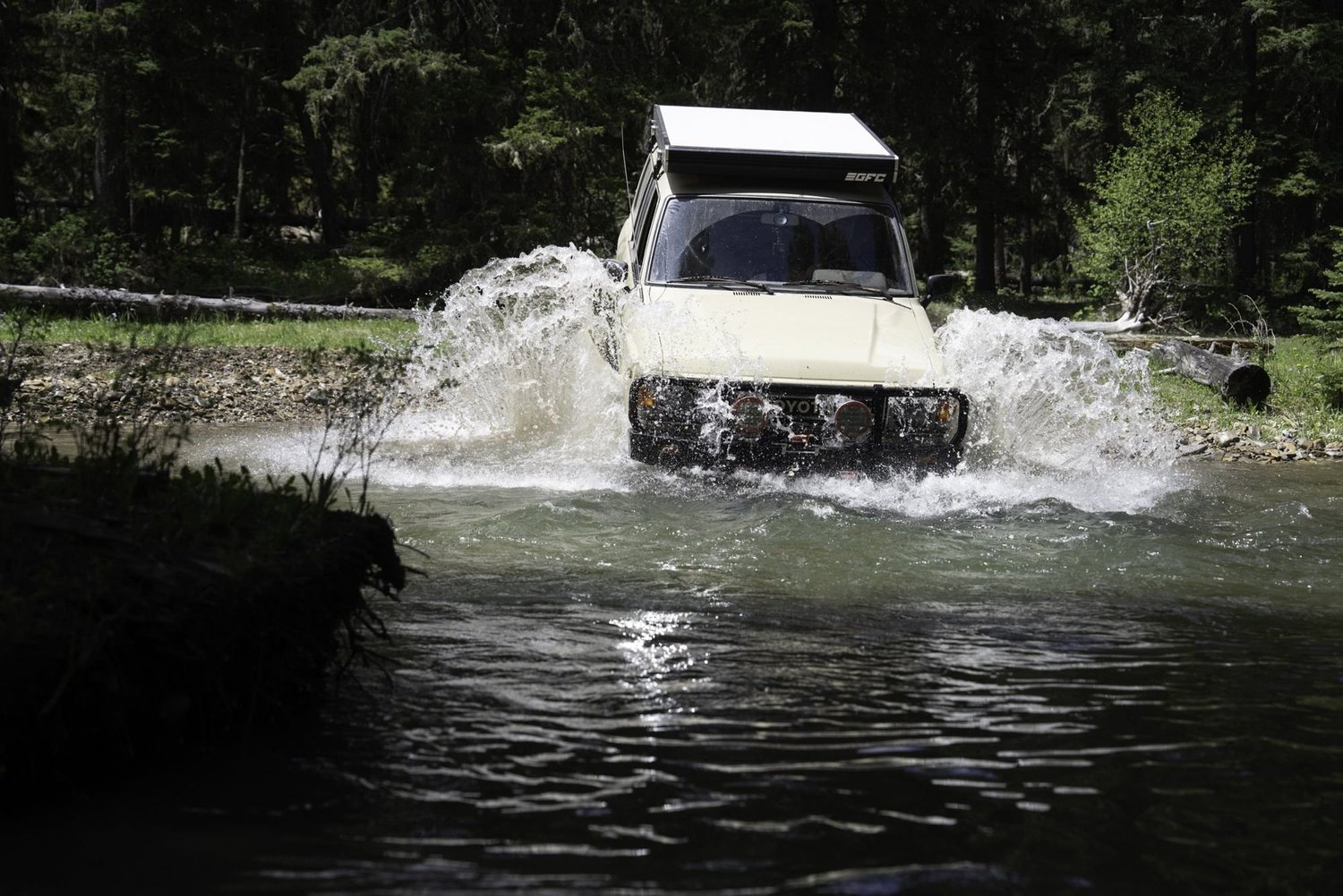 White truck with a rooftop tent throwing a huge splash in a dark river crossing on the Middle Fork Judith River road in the Little Belt Mountains, Montana