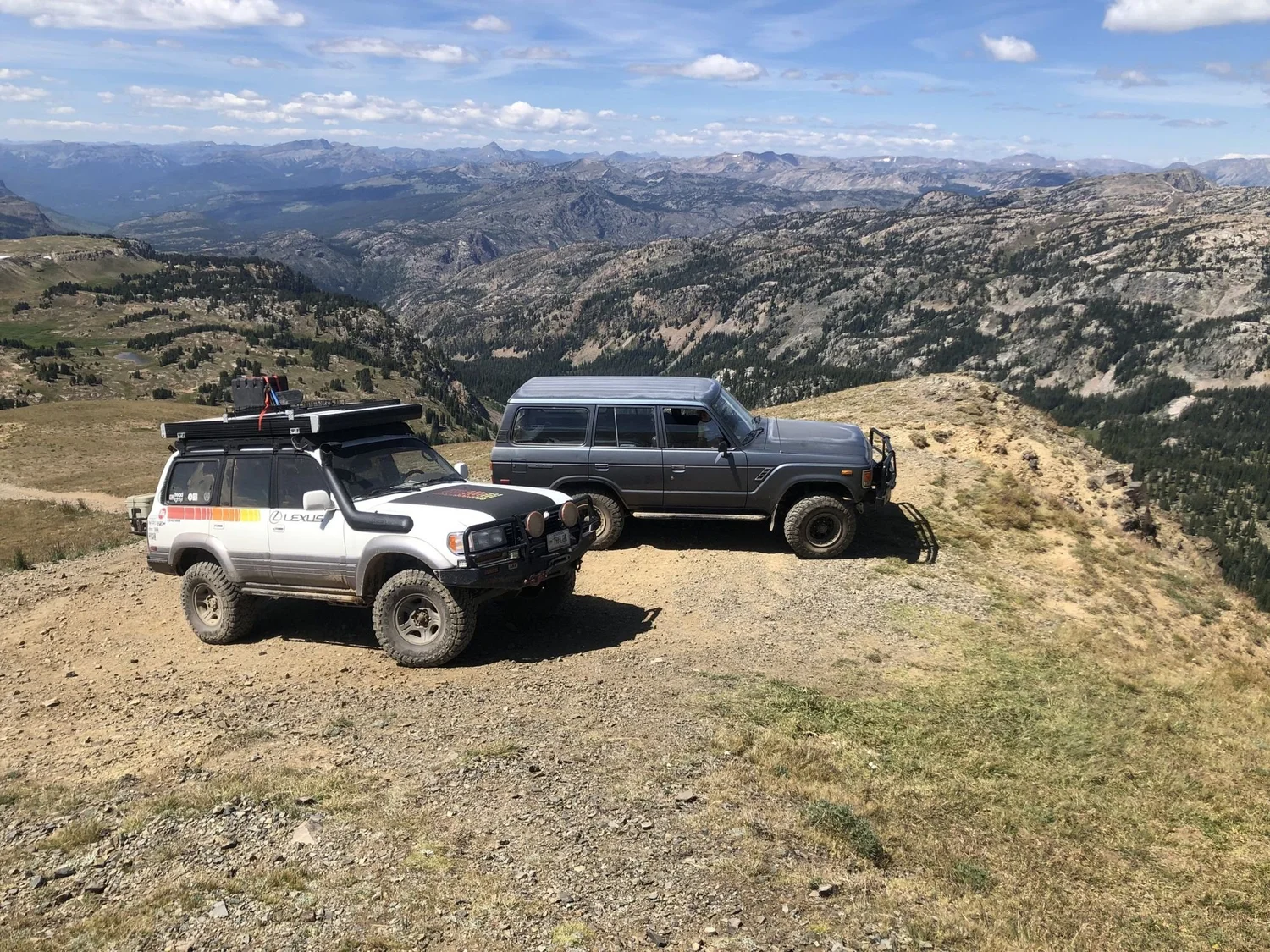 Striped Lexus LX450 and a gray Land Cruiser parked at a high overlook above a vast mountain basin near Cooke City, Montana