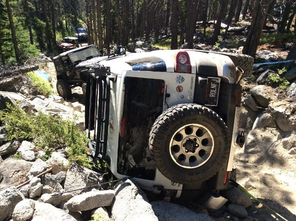 Rear view of a white FJ Cruiser lying on its side against boulders on a rocky forest trail in Montana